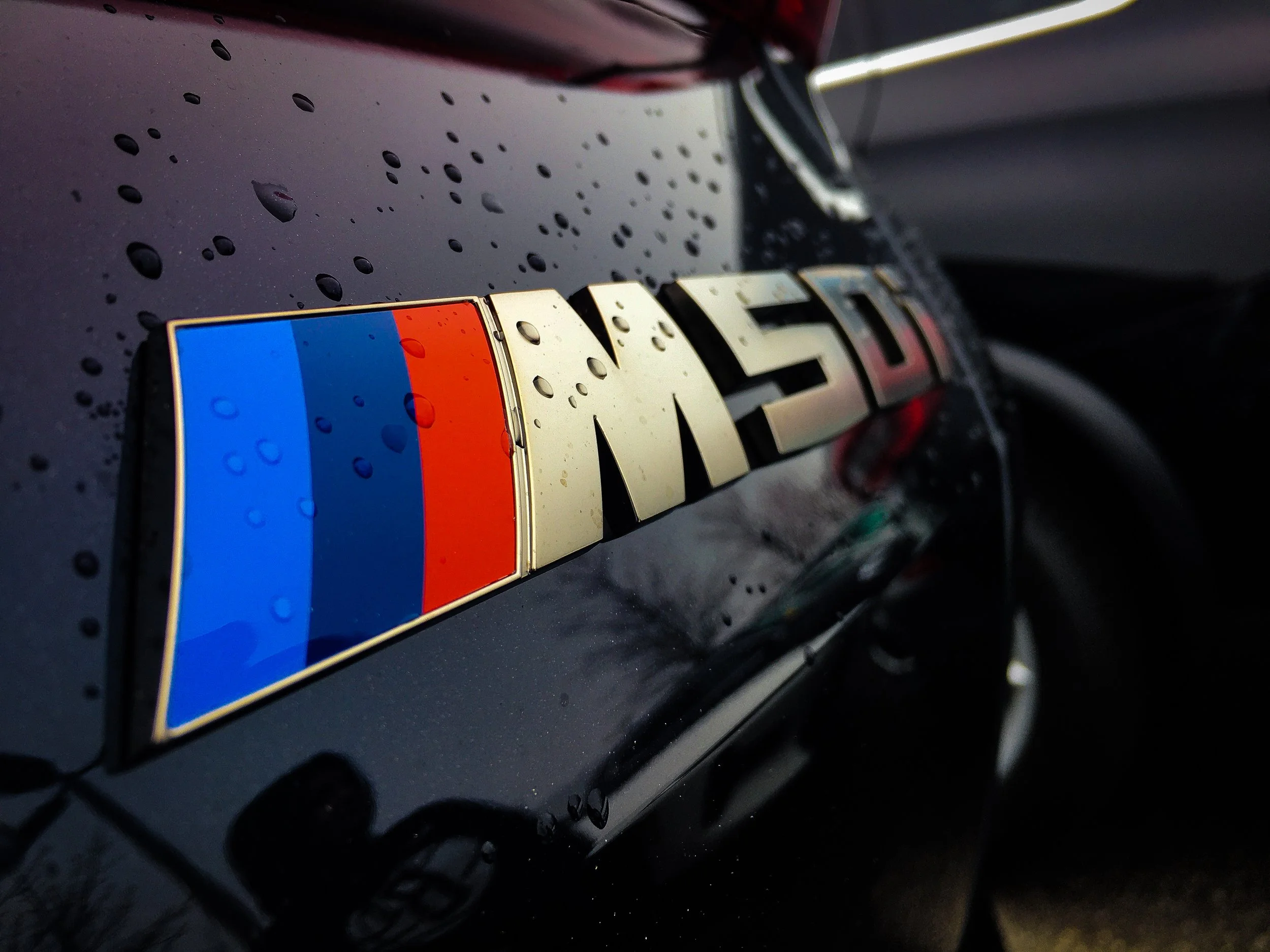Close-up of a car emblem with a multicolored stripe, water droplets on its surface, and a dark background.