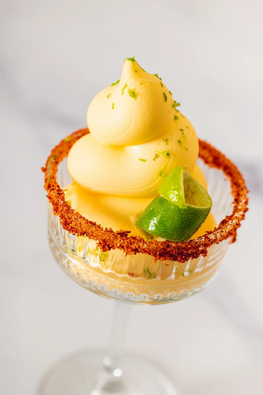 Lemon-themed dessert in a glass, topped with lemon-flavored whipped cream, garnished with lime zest and a lime slice, rimmed with red chili powder.
