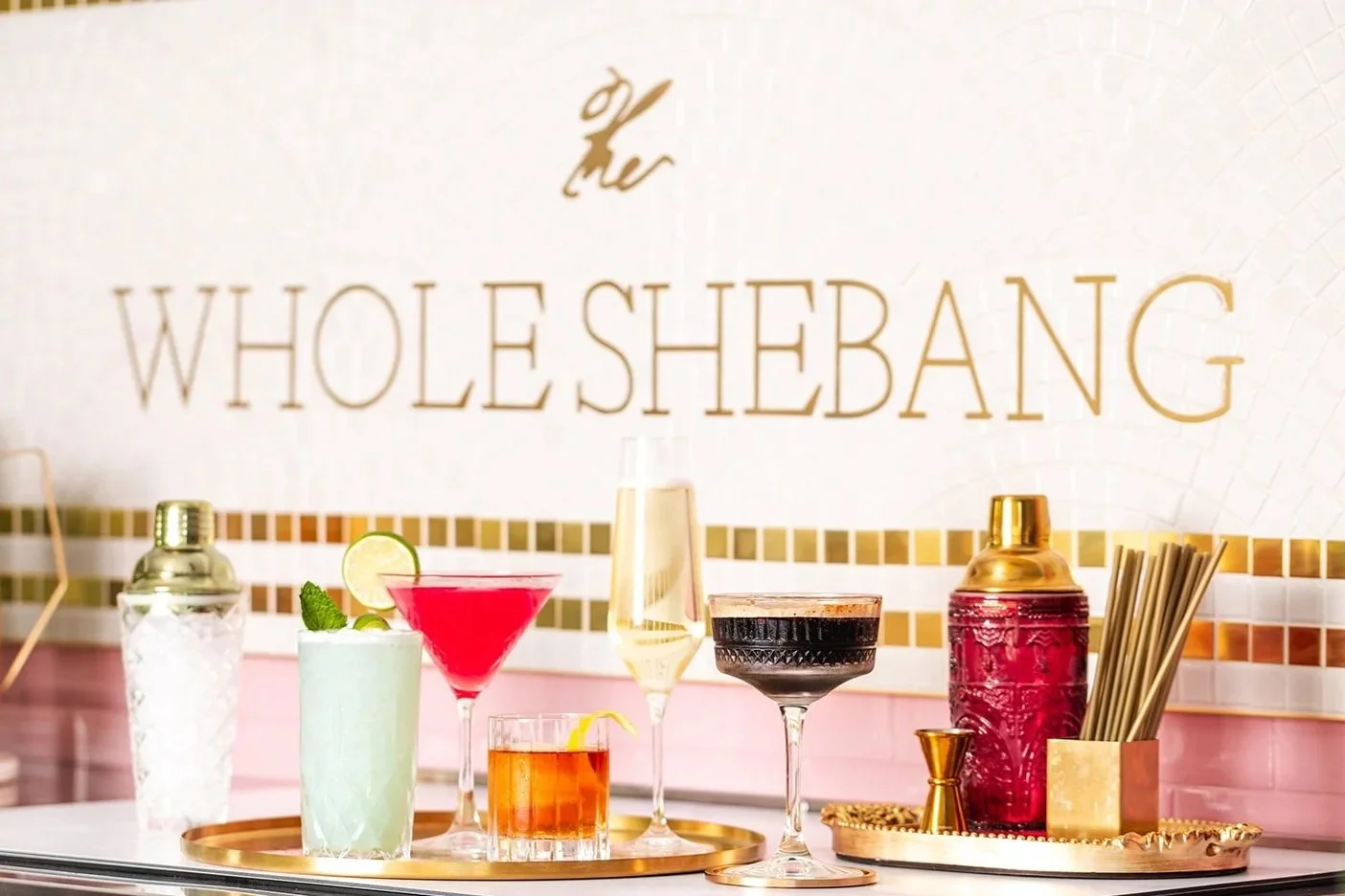 A variety of colorful cocktails on a tray in front of a sign that reads 'The Whole Shebang' with gold and pink accents.