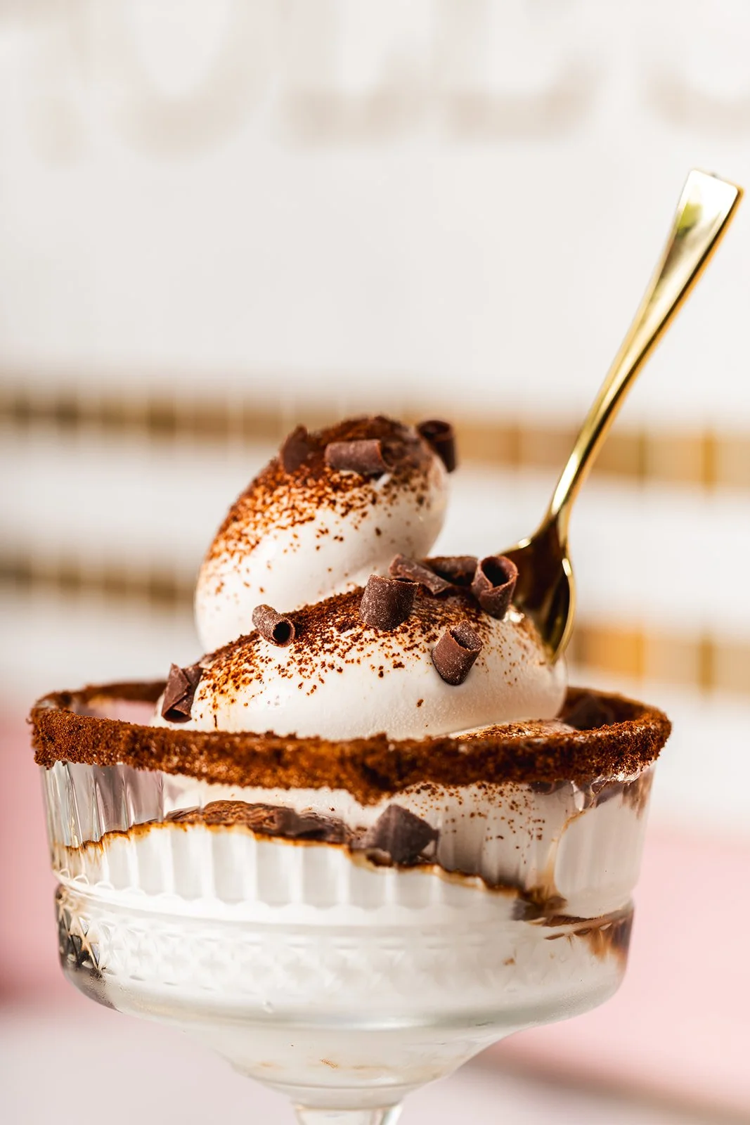 A glass dessert cup filled with a soft serve espresso martini, chocolate shavings, espresso dust, topped with scoops of ice cream and chocolate shavings, with a gold spoon.