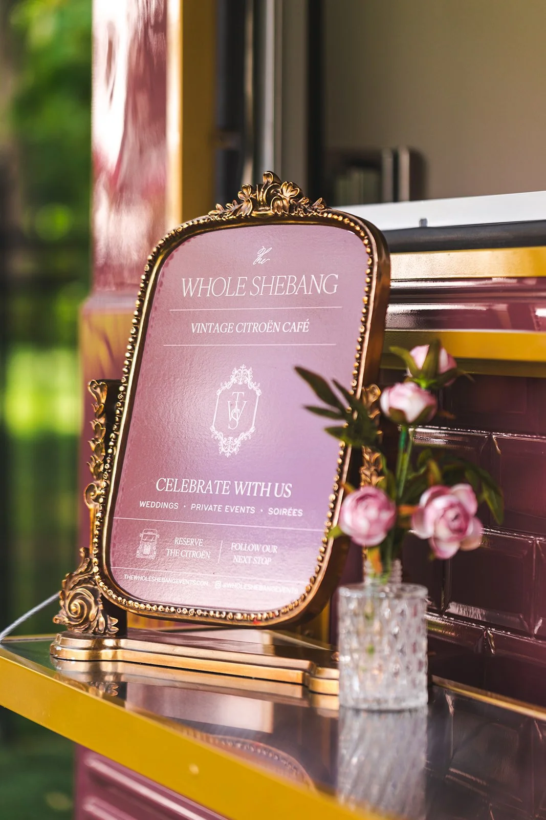 A decorative gold-framed sign on a yellow table promoting The Whole Shebang, a vintage Citroën café for weddings and private events, with a small pink flower arrangement in a glass vase beside it.