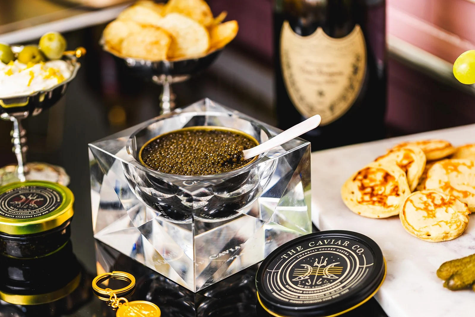 A bowl of black caviar with a white spoon inside, placed on a reflective, faceted glass display stand. Surrounding it are jars of caviar, plates of potato chips, and baked cheeses. In the background, there are bottles of champagne or wine and a marble serving surface.