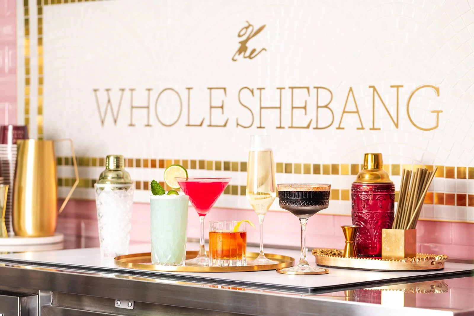 A display of colorful cocktails on a gold tray at a bar with a sign in the background reading 'The Whole Shebang.'