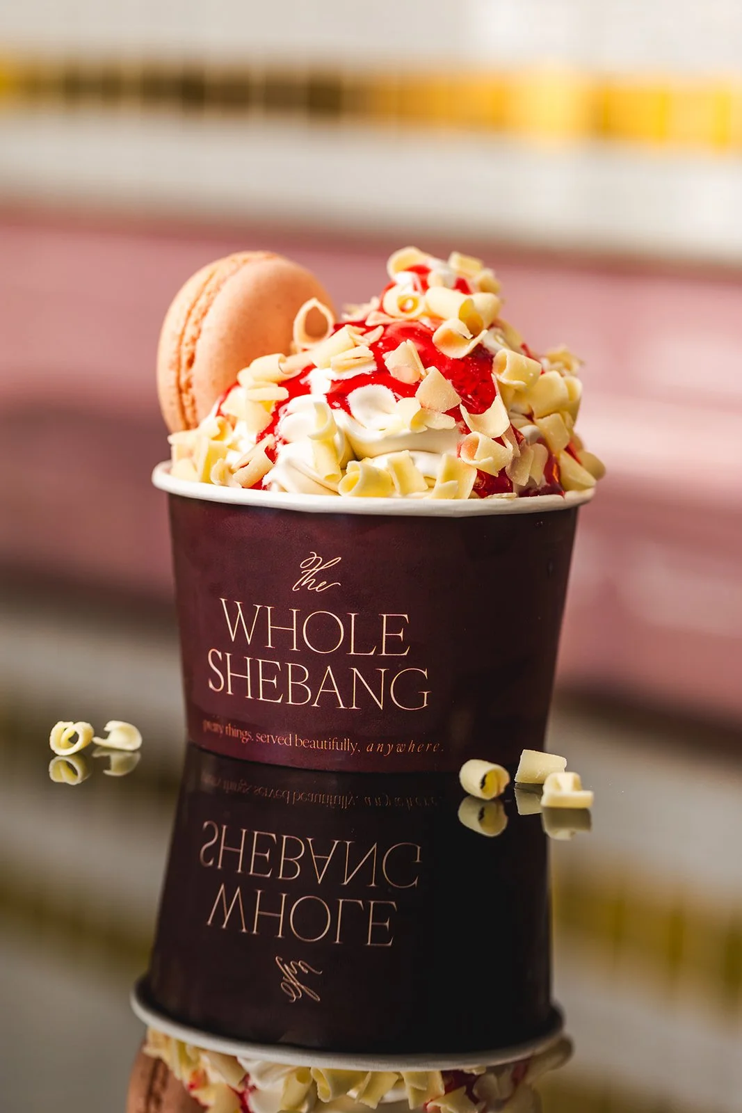 Cup of ice cream topped with white chocolate curls, strawberry syrup, a macaron, and white chocolate shavings, in a black paper cup with the logo 'The Whole Shebang' written on it.