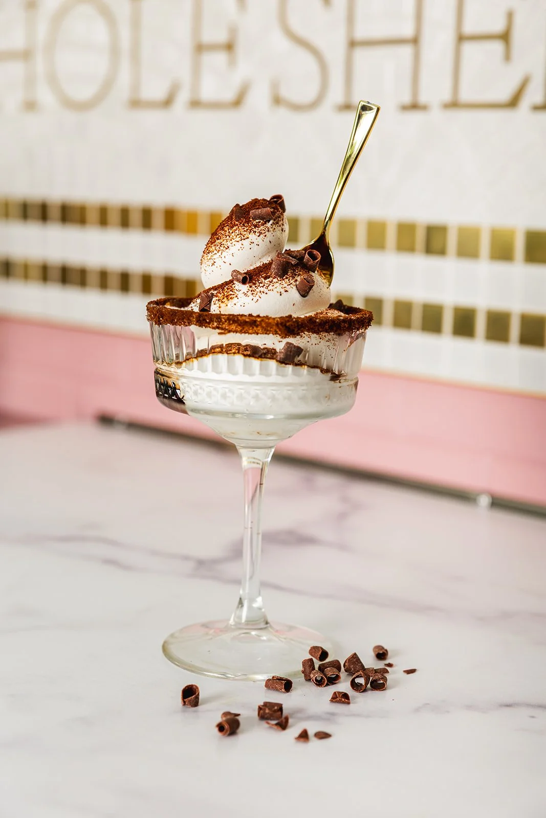 Soft Serve espresso martini in a glass bowl topped with whipped cream, chocolate shavings, and cocoa powder, with a gold spoon inside.