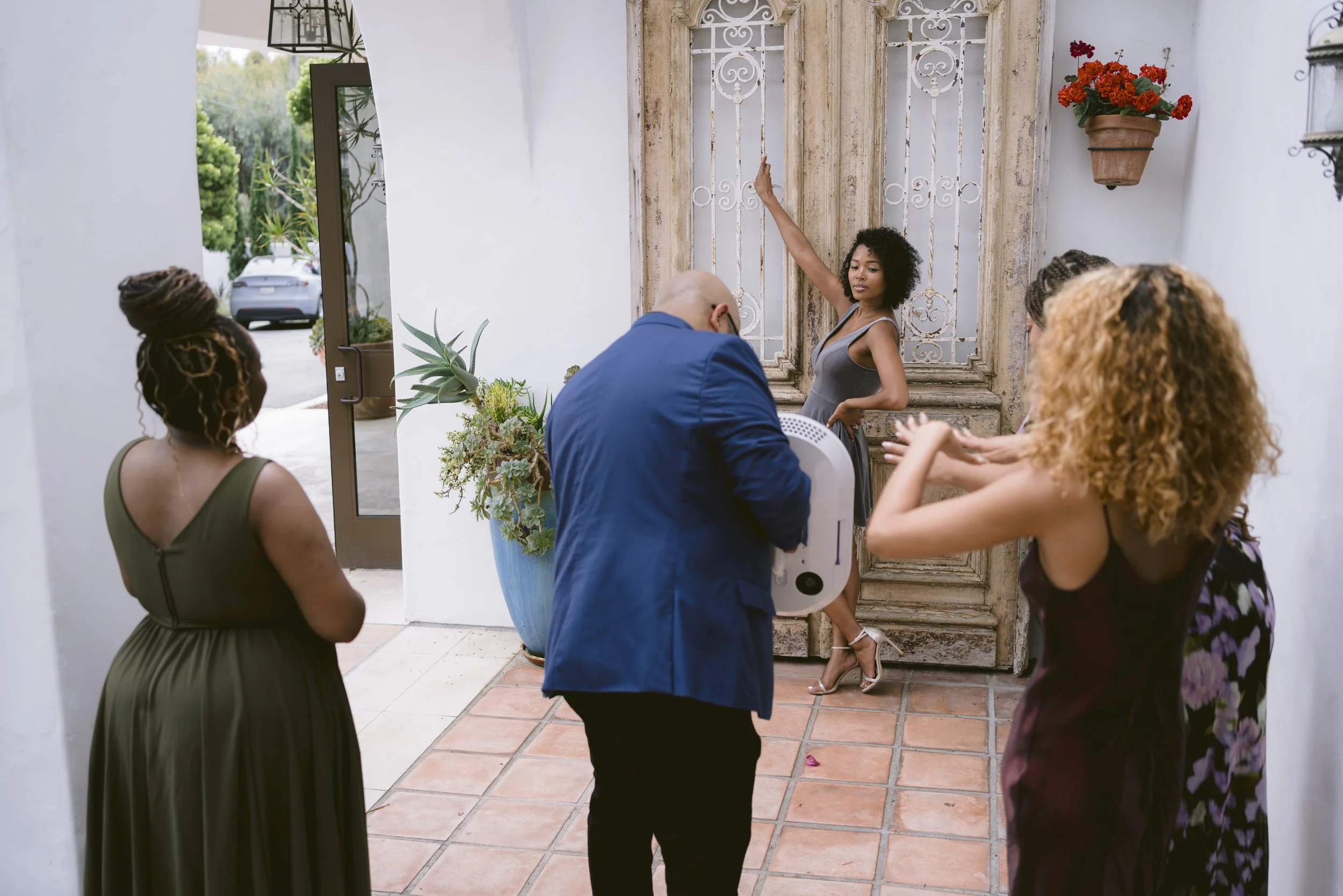 A photoshoot scene with a woman posing near an ornate window, surrounded by four women and a photographer, outside a white building with potted plants and a car in the background.