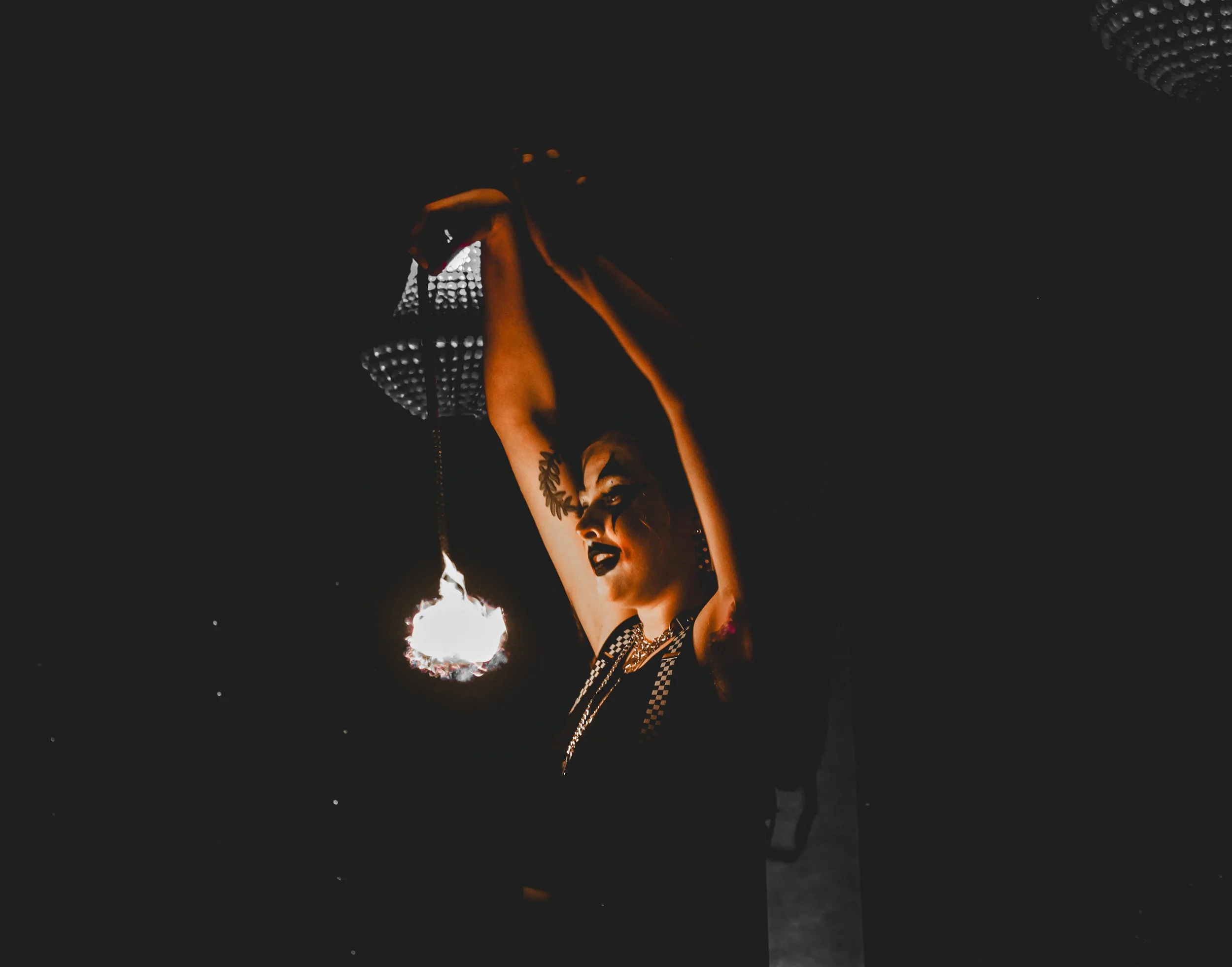 A woman with tattoos and dark makeup holds a flaming object in a dark environment with a reflective ceiling.