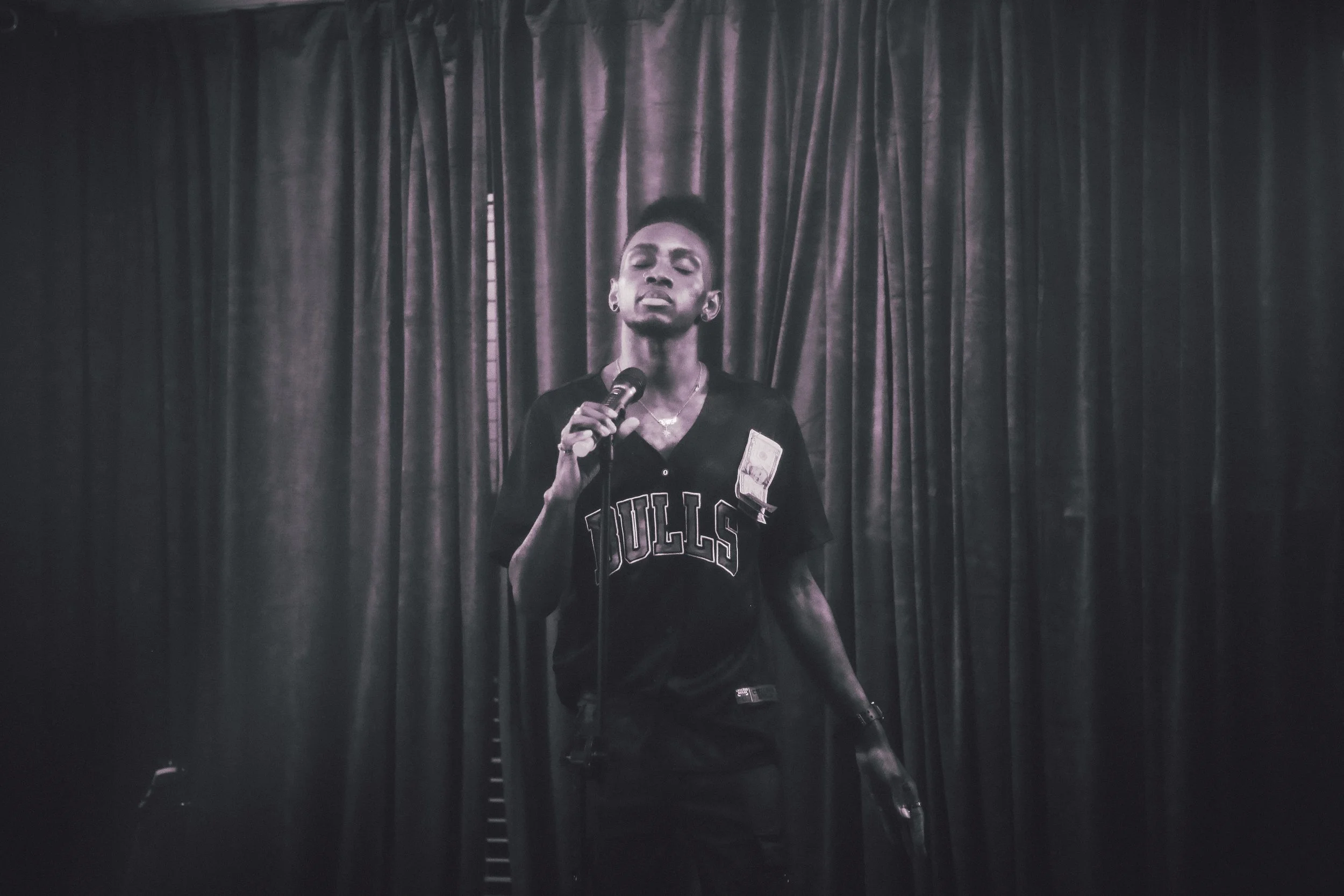 A person with short hair standing in front of a dark curtain, holding a microphone close to their mouth, wearing a Chicago Bulls jersey, with a dollar bill pinned to their chest.