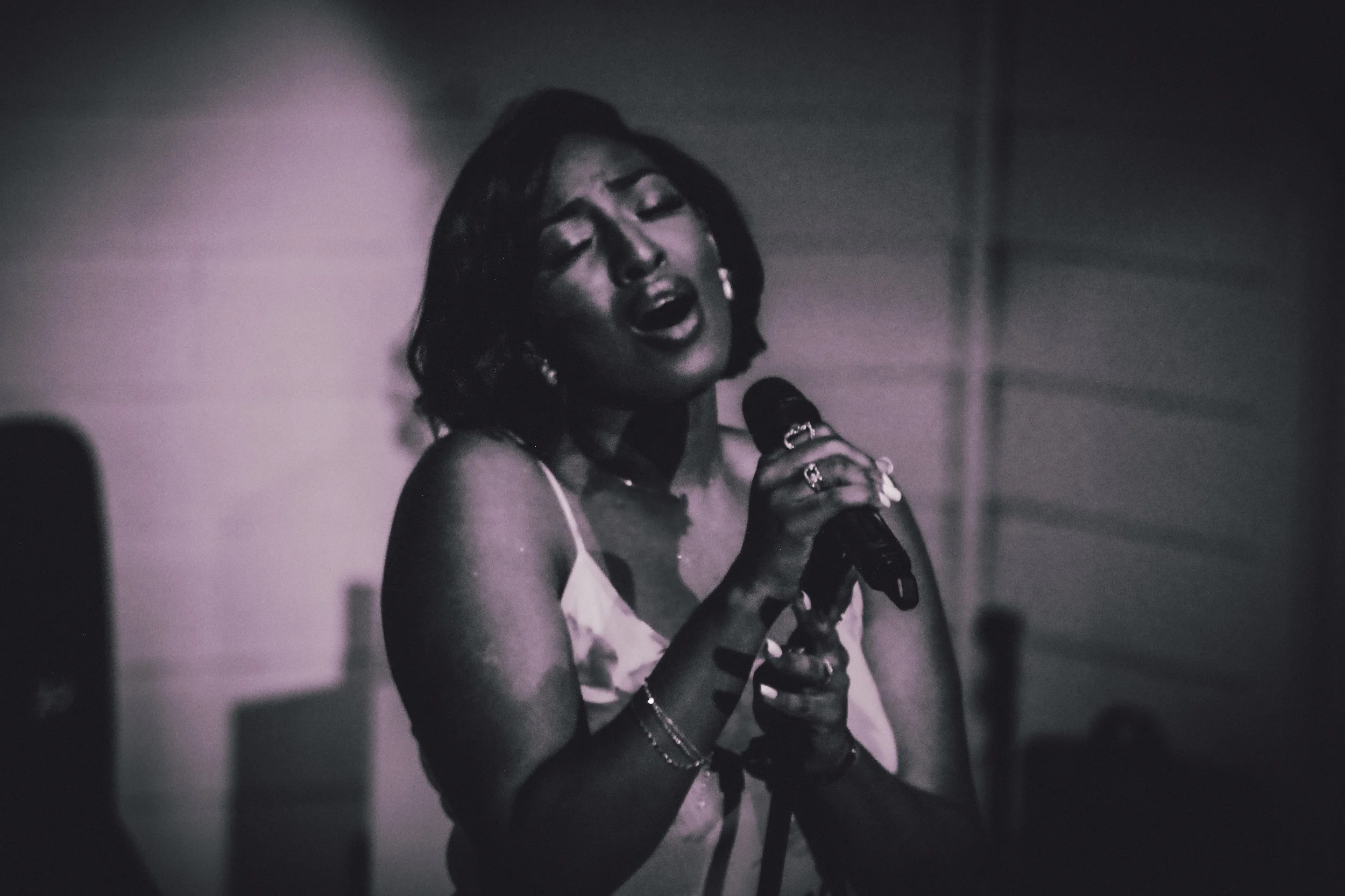 A woman singing into a microphone with her eyes closed, wearing a sleeveless top, rings, and bracelets, in a black and white photo.