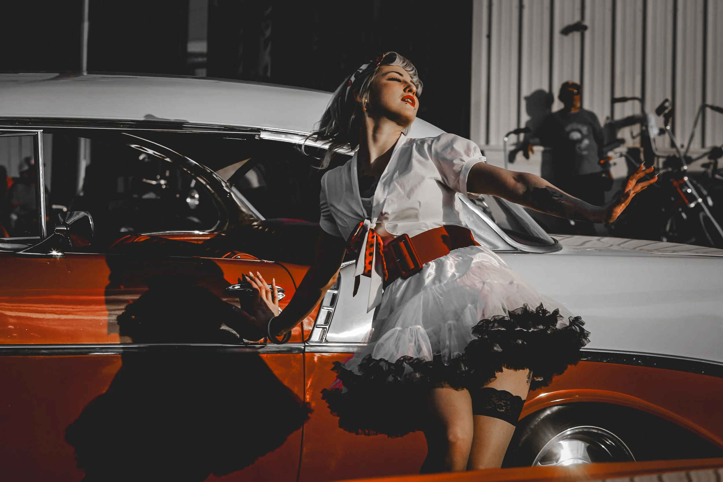 A woman with vintage makeup and blonde hair styled in retro waves, wearing a white blouse with a red and black sash, a black lace garter, and a black petticoat, is posing on an orange and white classic car with her right hand on the car's door and he