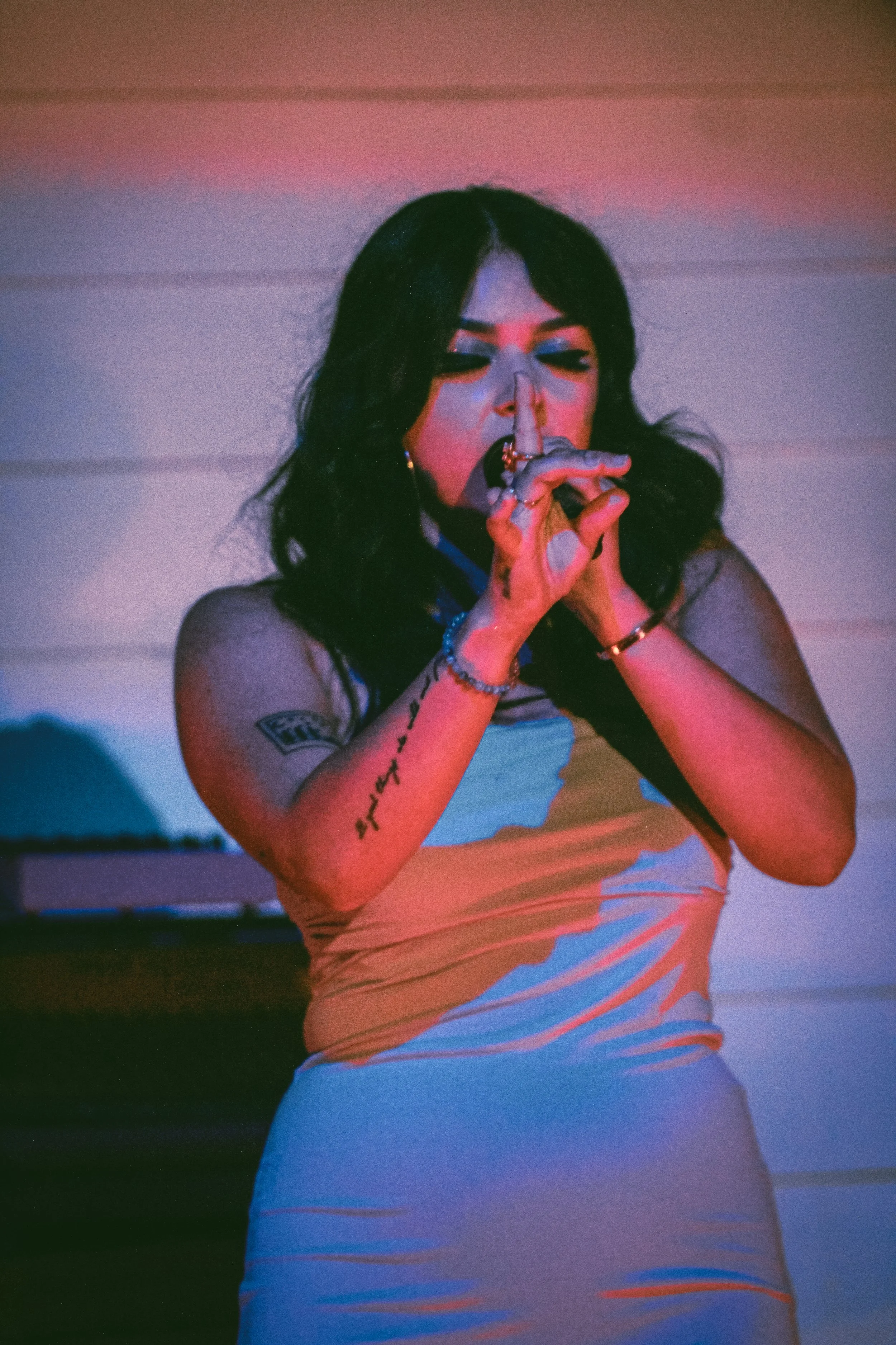 A woman with black hair and tattoos on her left arm is wearing a sleeveless top and making a finger gun gesture with her right hand near her lips, with a colorful background of pink and blue hues.