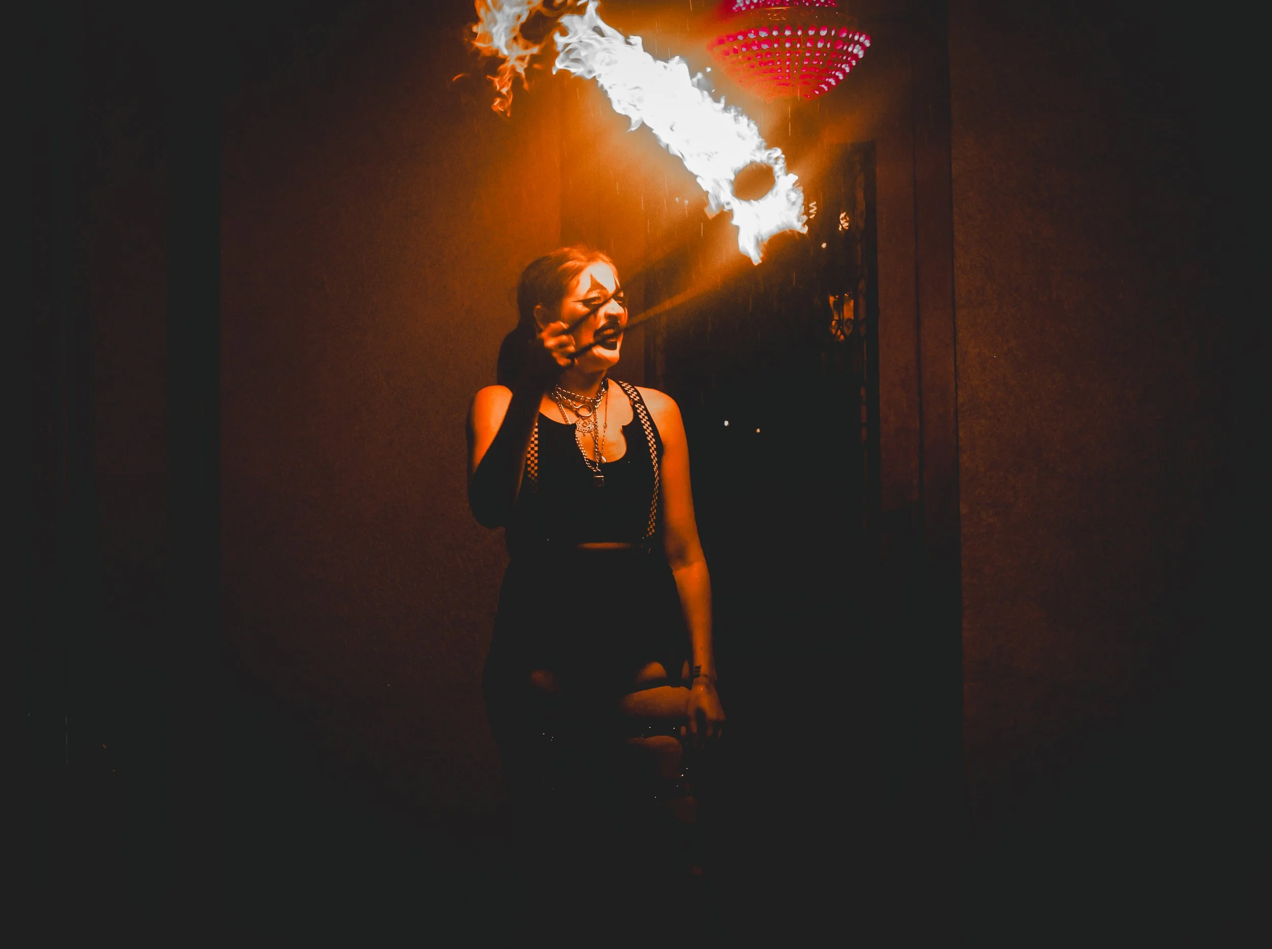 A woman in black clothing with chains, holding a lit torch that is producing a flame and smoke, standing in a dimly lit indoor space.