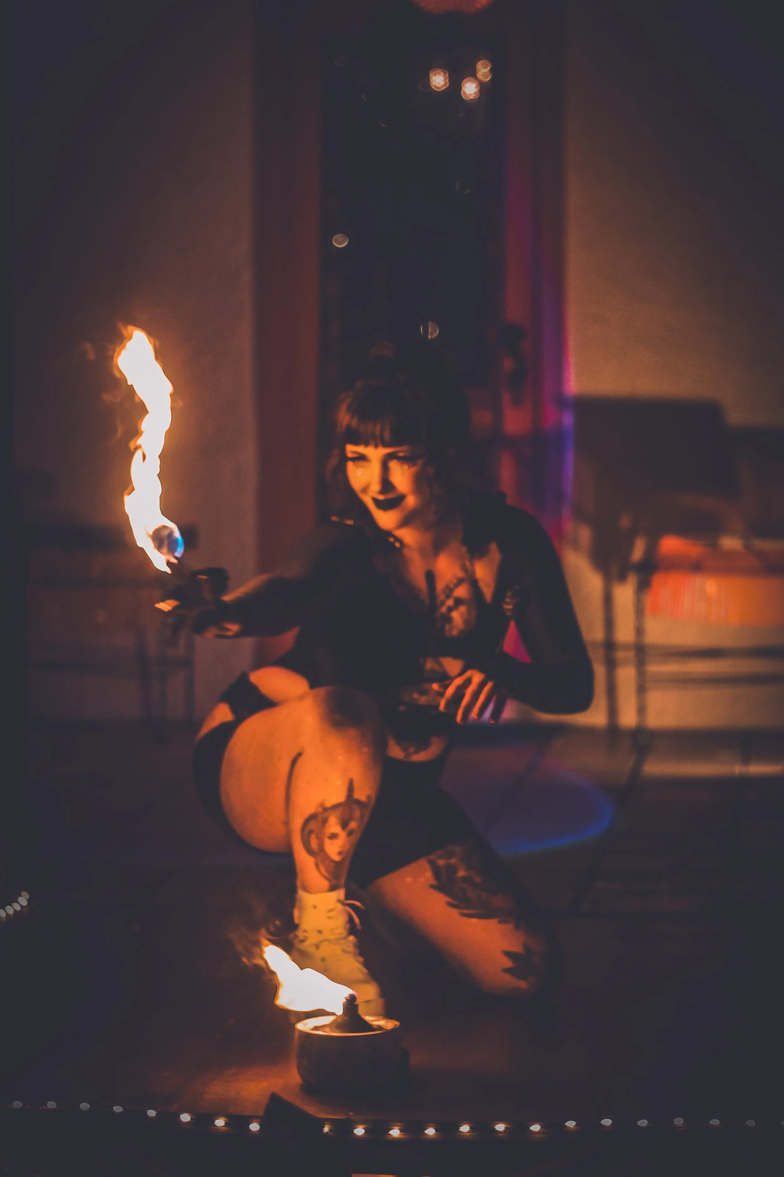 A woman with tattoos and black hair performing a fire dance in a dimly lit room, holding a flaming torch and kneeling near a fire bowl.