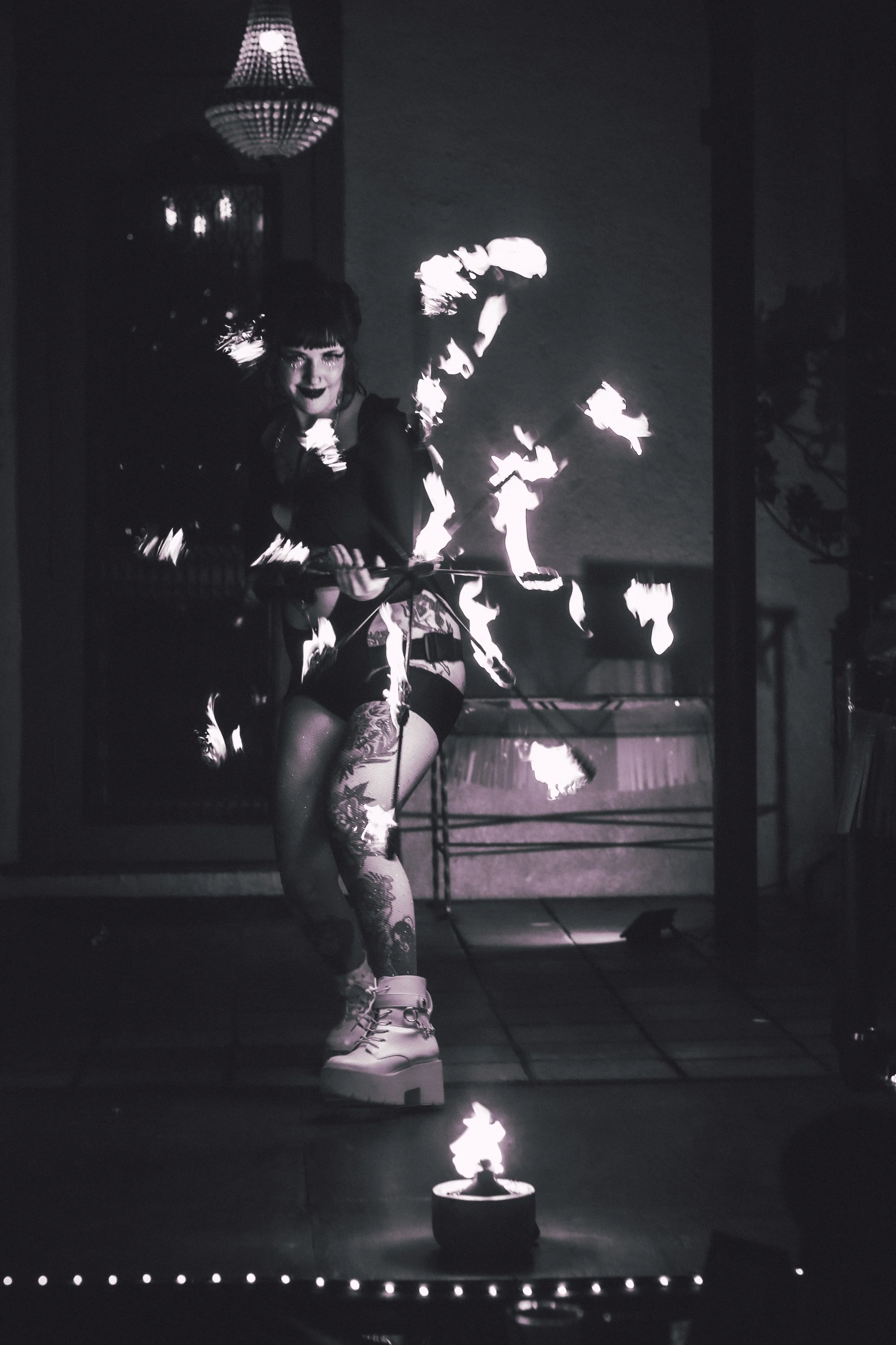 A woman performs fire juggling indoors, surrounded by flames, with a chandelier above her.