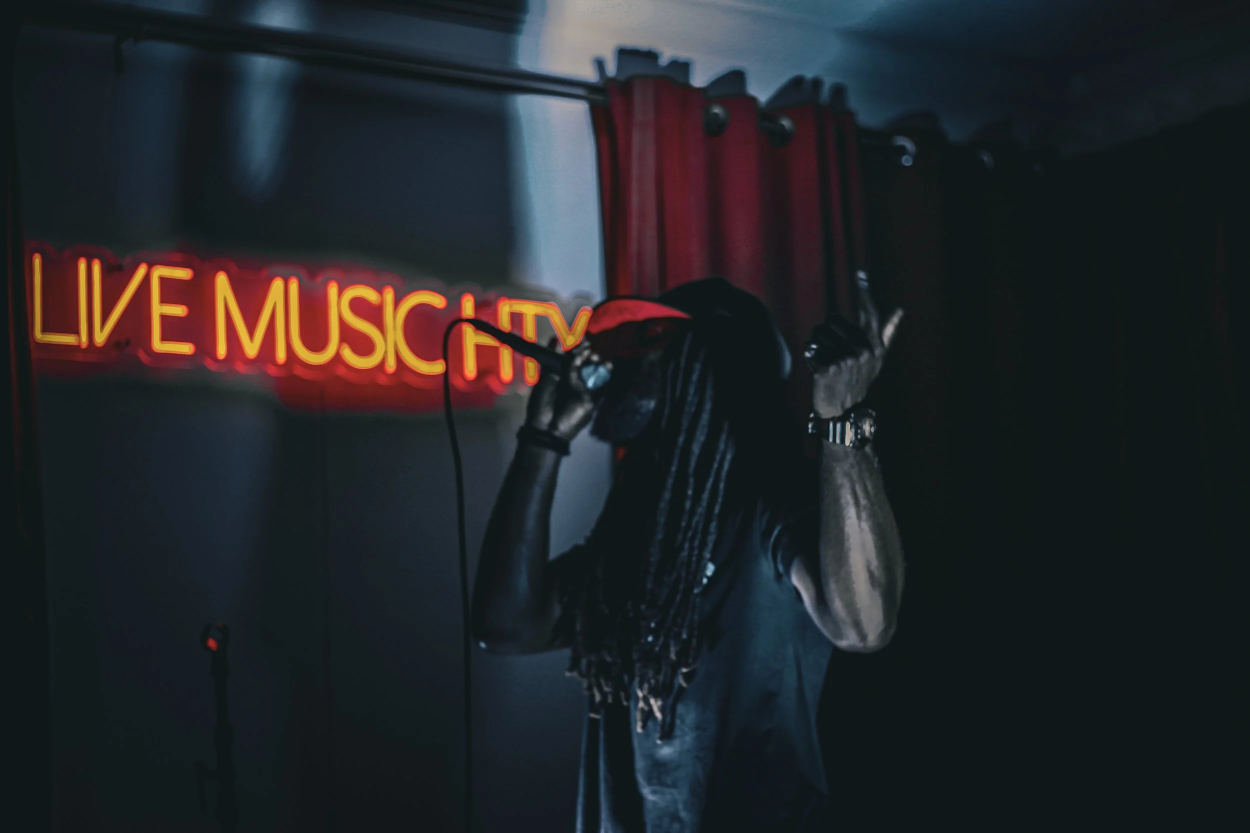 A person with long dreadlocks wearing headphones and a red cap, adjusting the headphones in a dimly lit room with a neon sign reading 'LIVE MUSIC TV' in the background.