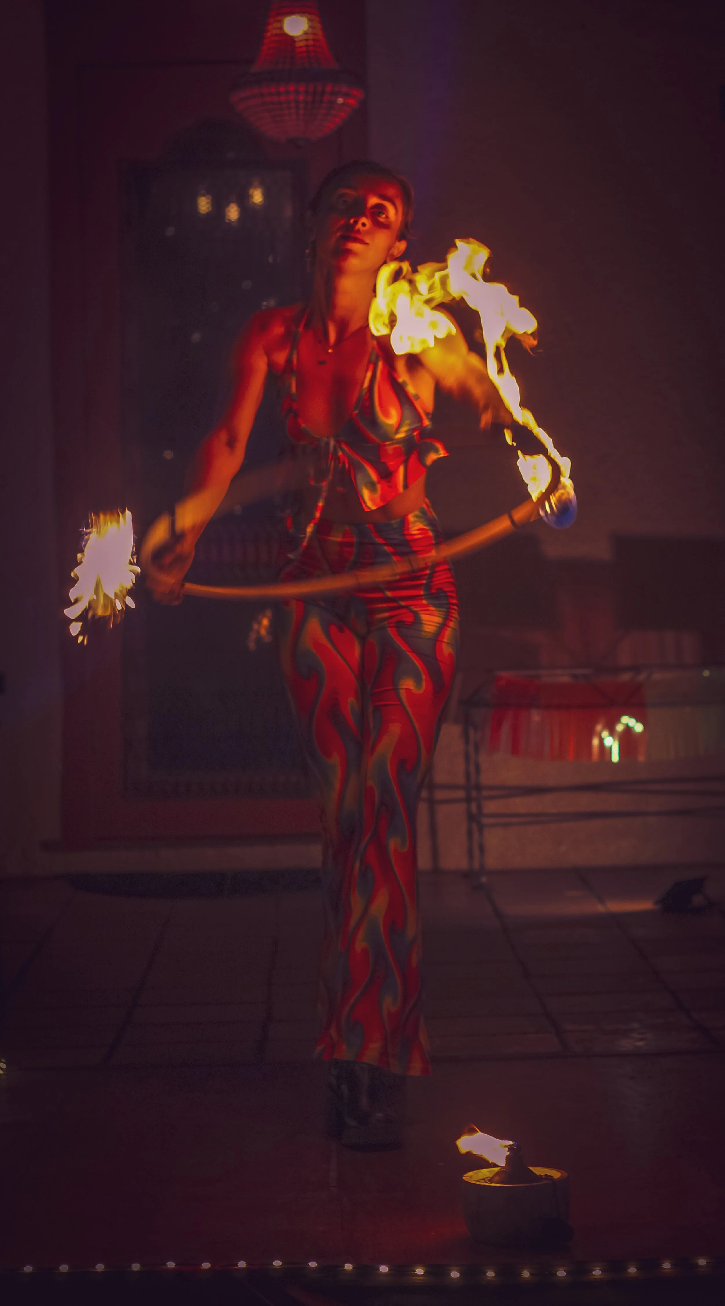 A female fire performer dressed in colorful flame-patterned clothing, spinning fire poi in an indoor setting.
