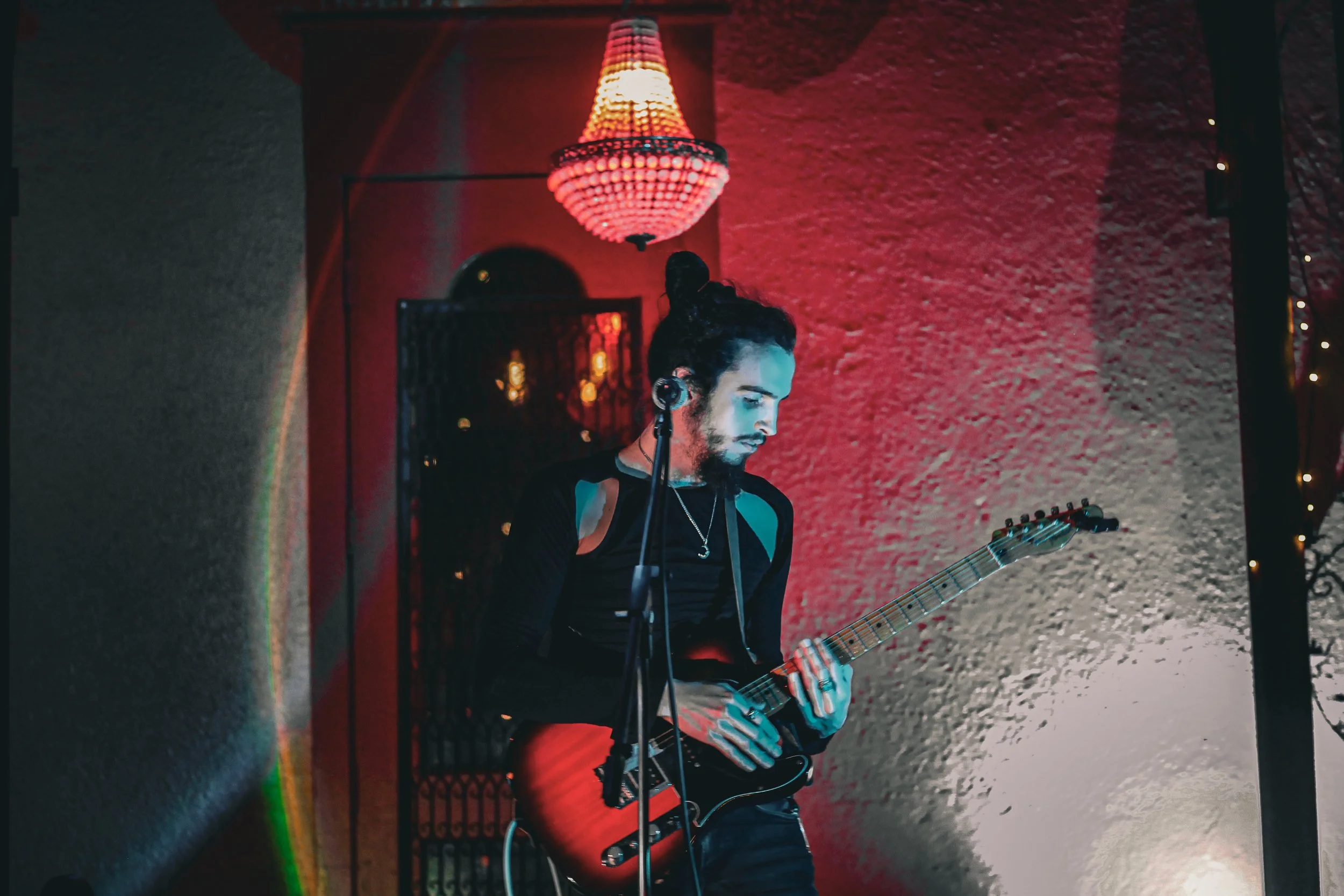 A musician with long dark hair tied back, wearing a black top, playing an electric guitar indoors under red and warm lighting.
