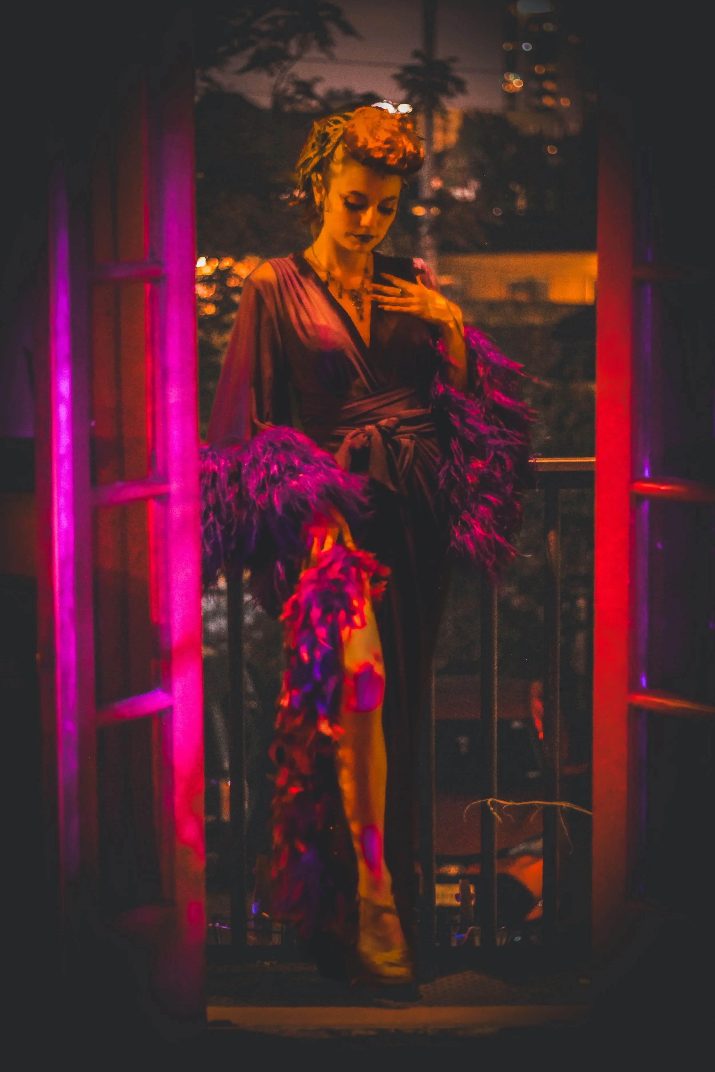 A woman on a balcony at night, dressed in a satin gown with feathered sleeves, looking at her phone with one hand on her chest, illuminated by pink and purple lighting.