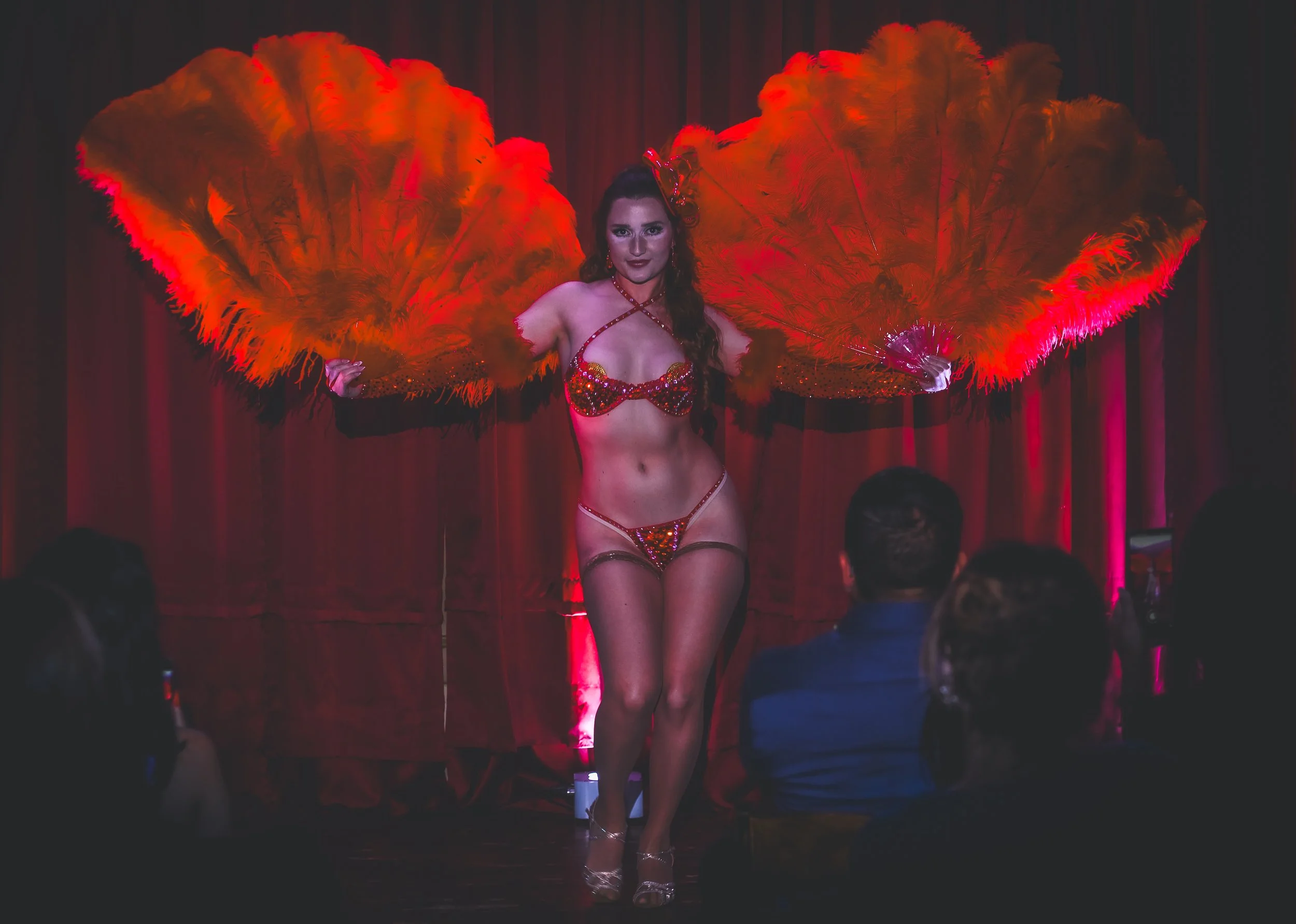 A woman performing on stage wearing a red sequin bikini and holding large orange feather fans with red lighting and an audience watching