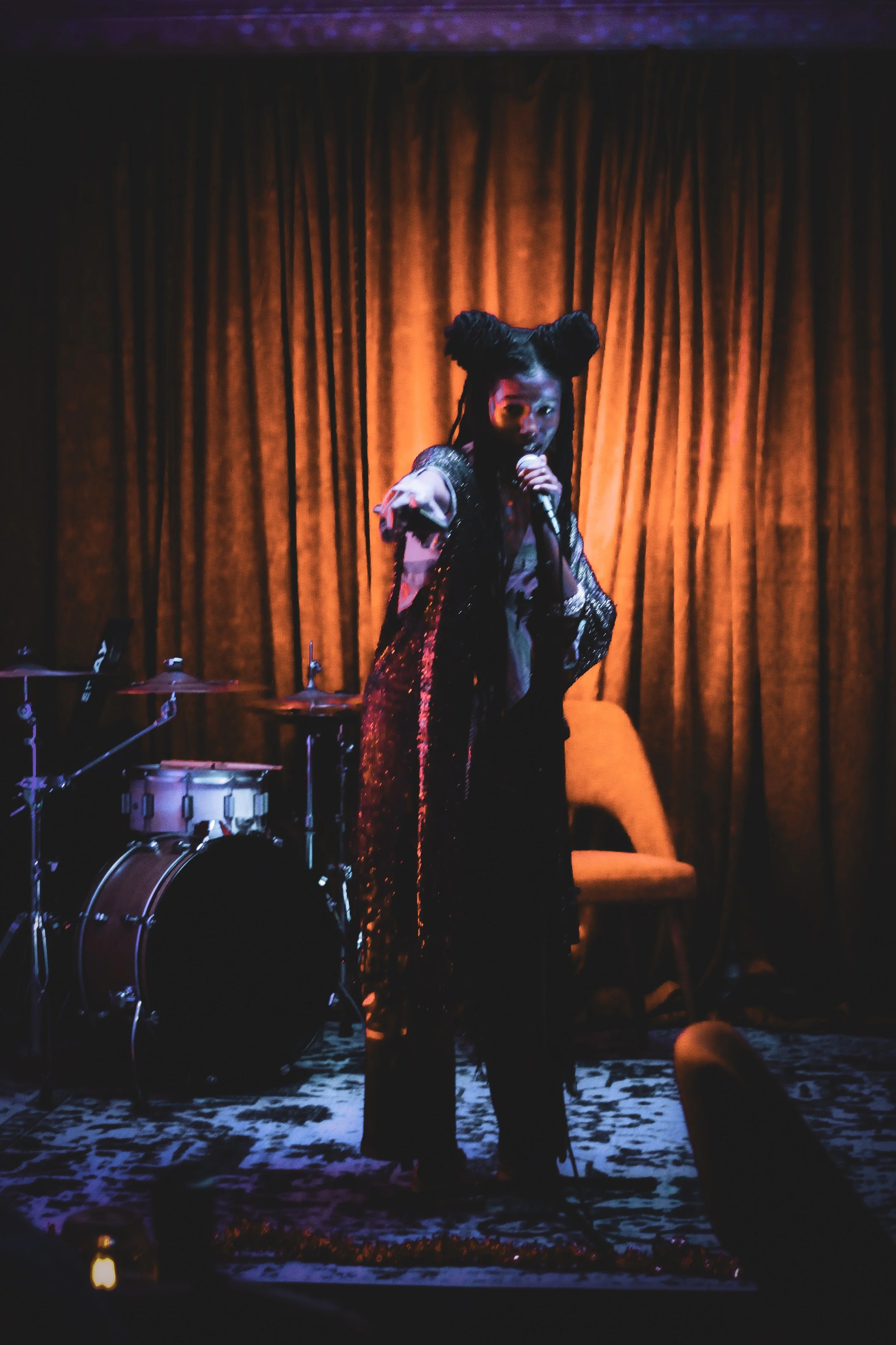 Performer on stage singing into a microphone with a drum set and a chair in the background, illuminated by warm stage lighting.