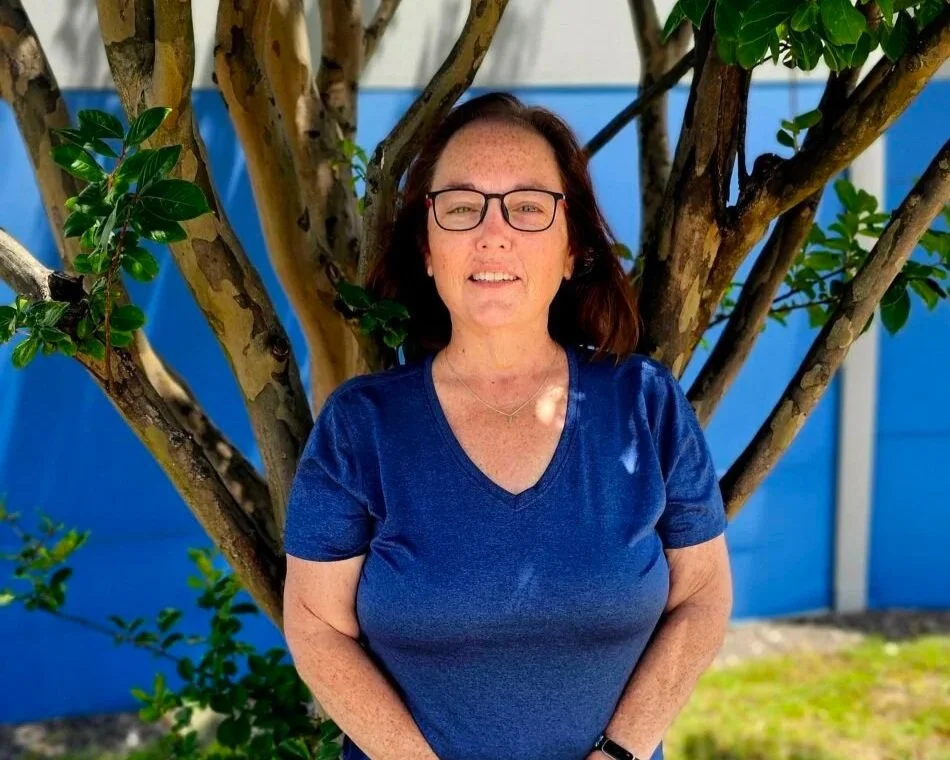 A woman with glasses and freckles wearing a blue shirt stands outdoors in front of a tree with green leaves and a blue fence in the background.