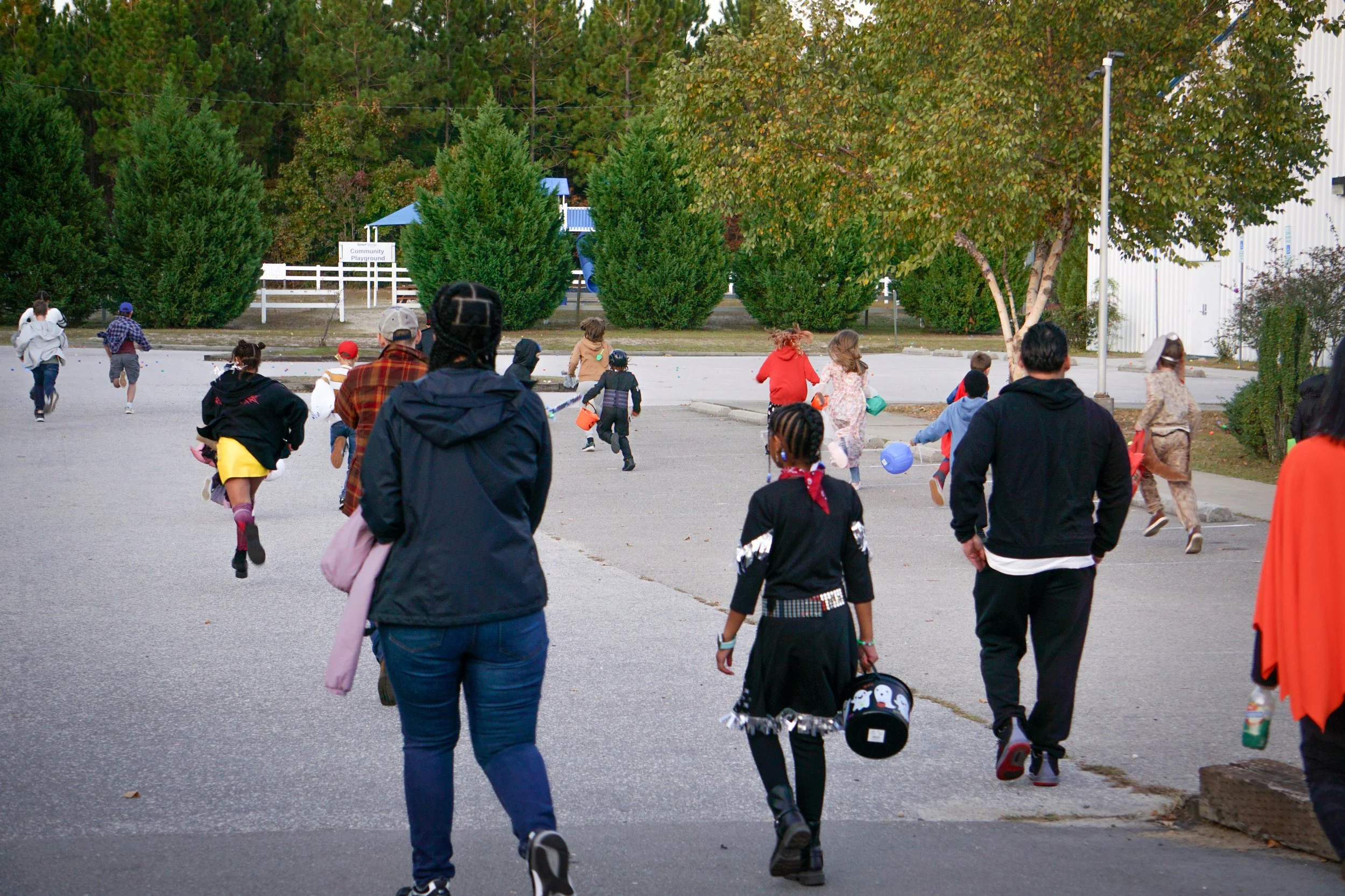 Children and adults running towards a community playground area with trees and a white fence in the background.
