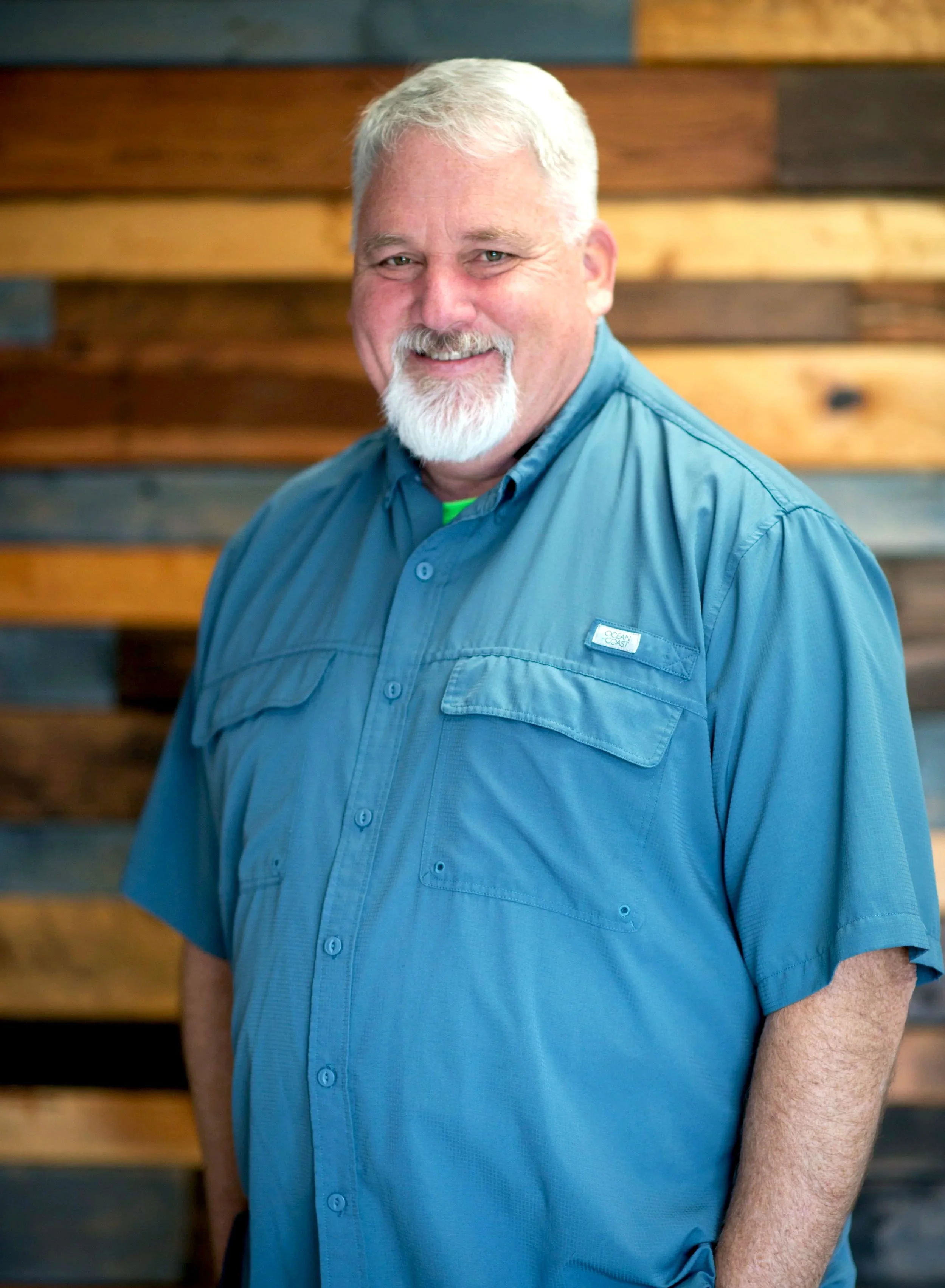 A smiling man with white hair and a beard wearing a blue short-sleeved button-up shirt standing in front of a wooden wall background.