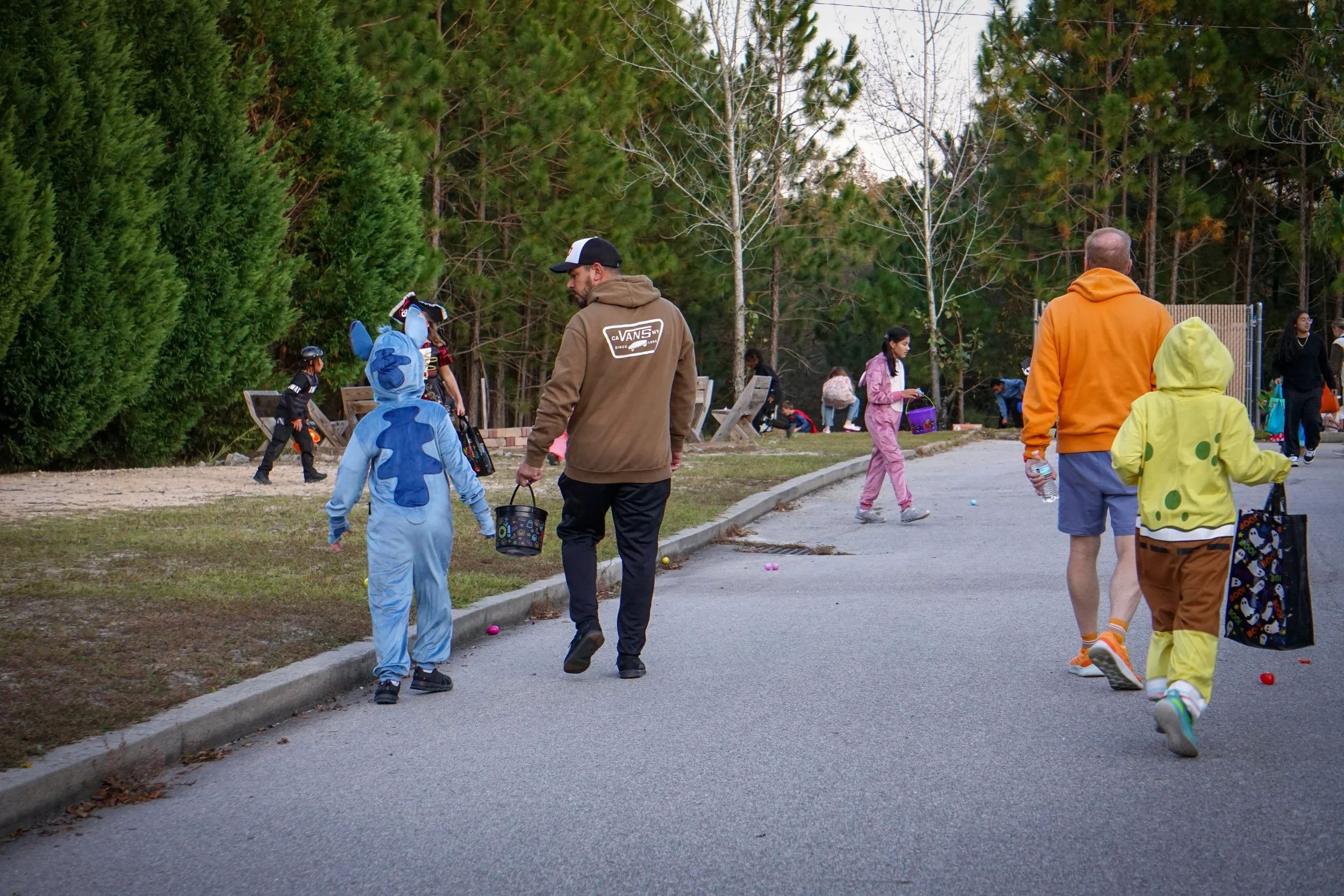 People celebrating Halloween with children in costumes, walking outdoors among trees, with some holding buckets for collecting candy.