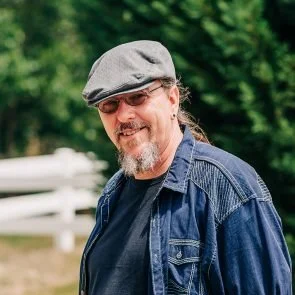 A man with a beard and glasses wearing a gray cap and denim jacket outdoors with green trees in the background.