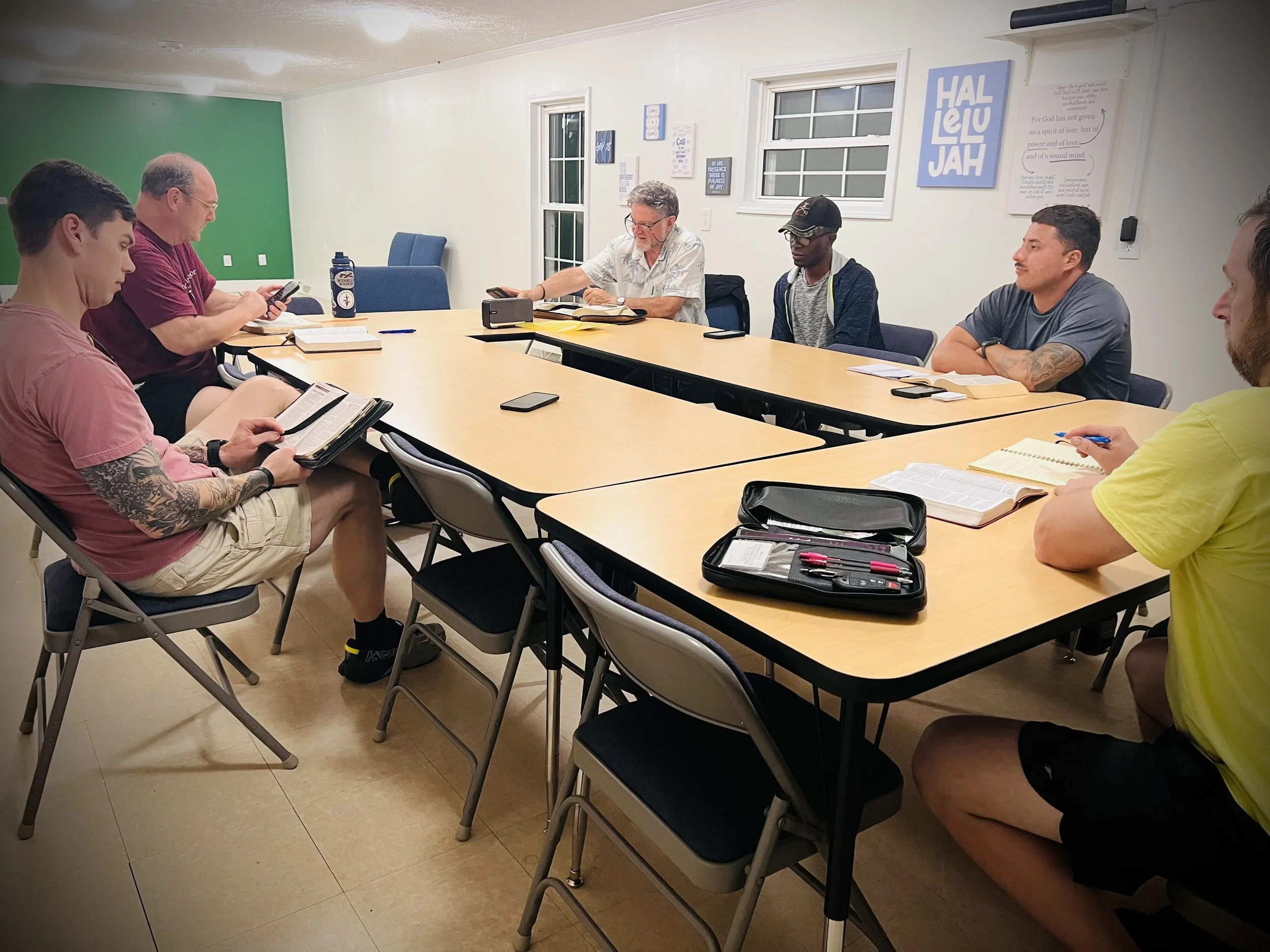 Six men sitting around a rectangular conference table in a room, some are reading or looking at their phones, with a variety of books, notebooks, and pens on the table. The room has white walls with some framed artworks and phrases, and a green accent wall in the background.