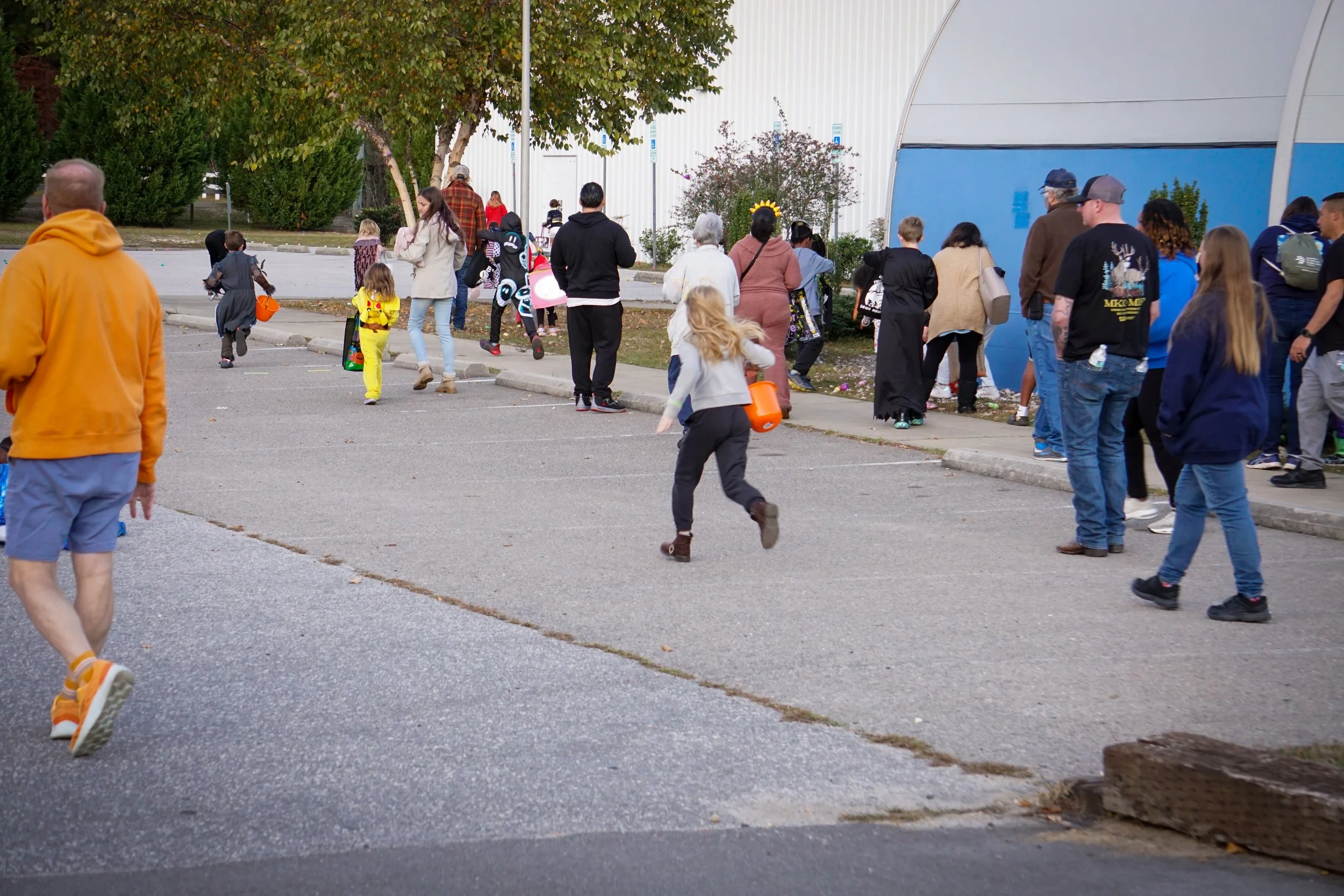 People walking and gathering outdoors near a building with blue and white walls, some carrying buckets and wearing Halloween costumes, during what appears to be a Halloween event.