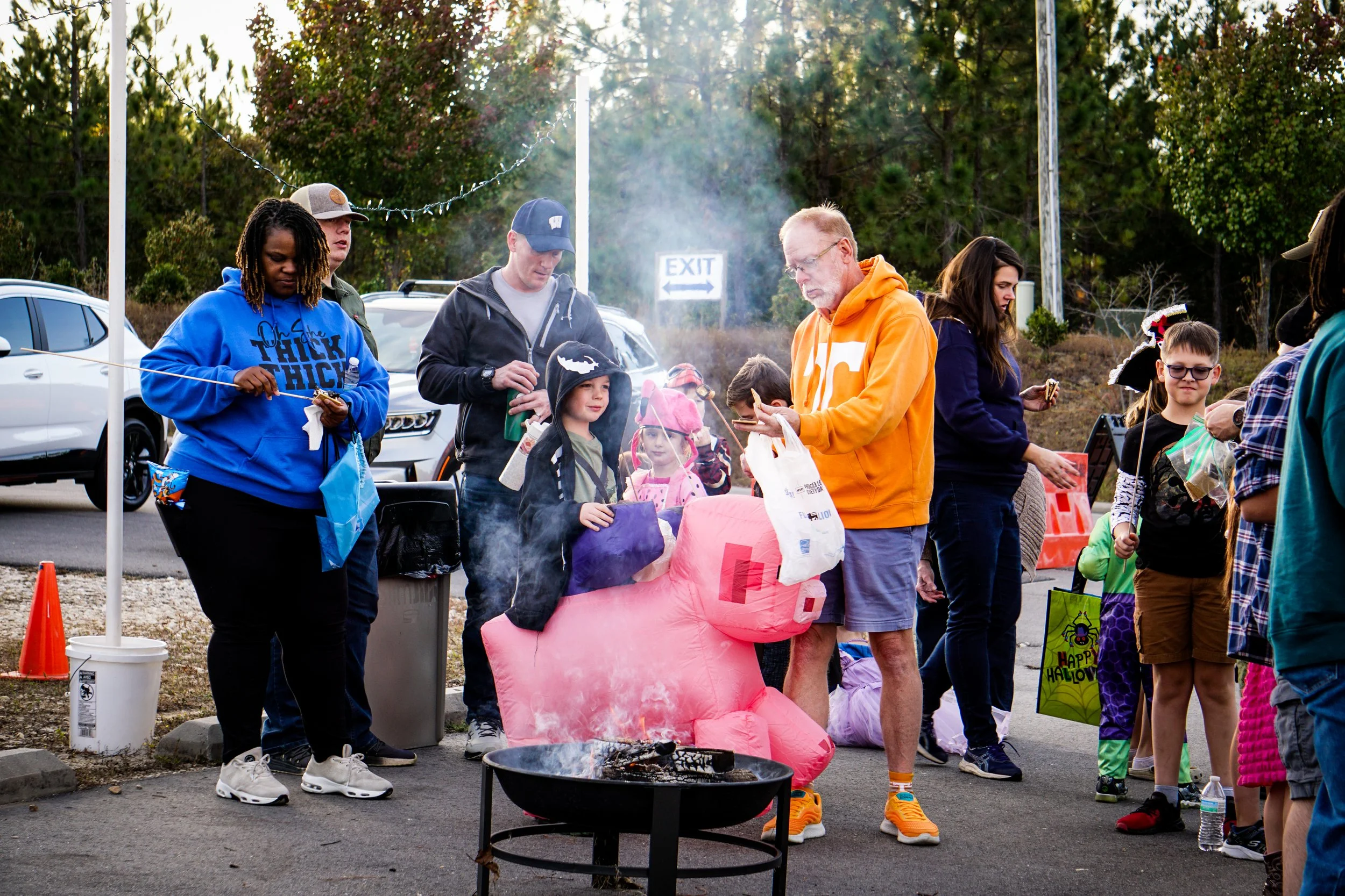People gathered around a bonfire at an outdoor Halloween event with children in costumes and an 'EXIT' sign in the background.