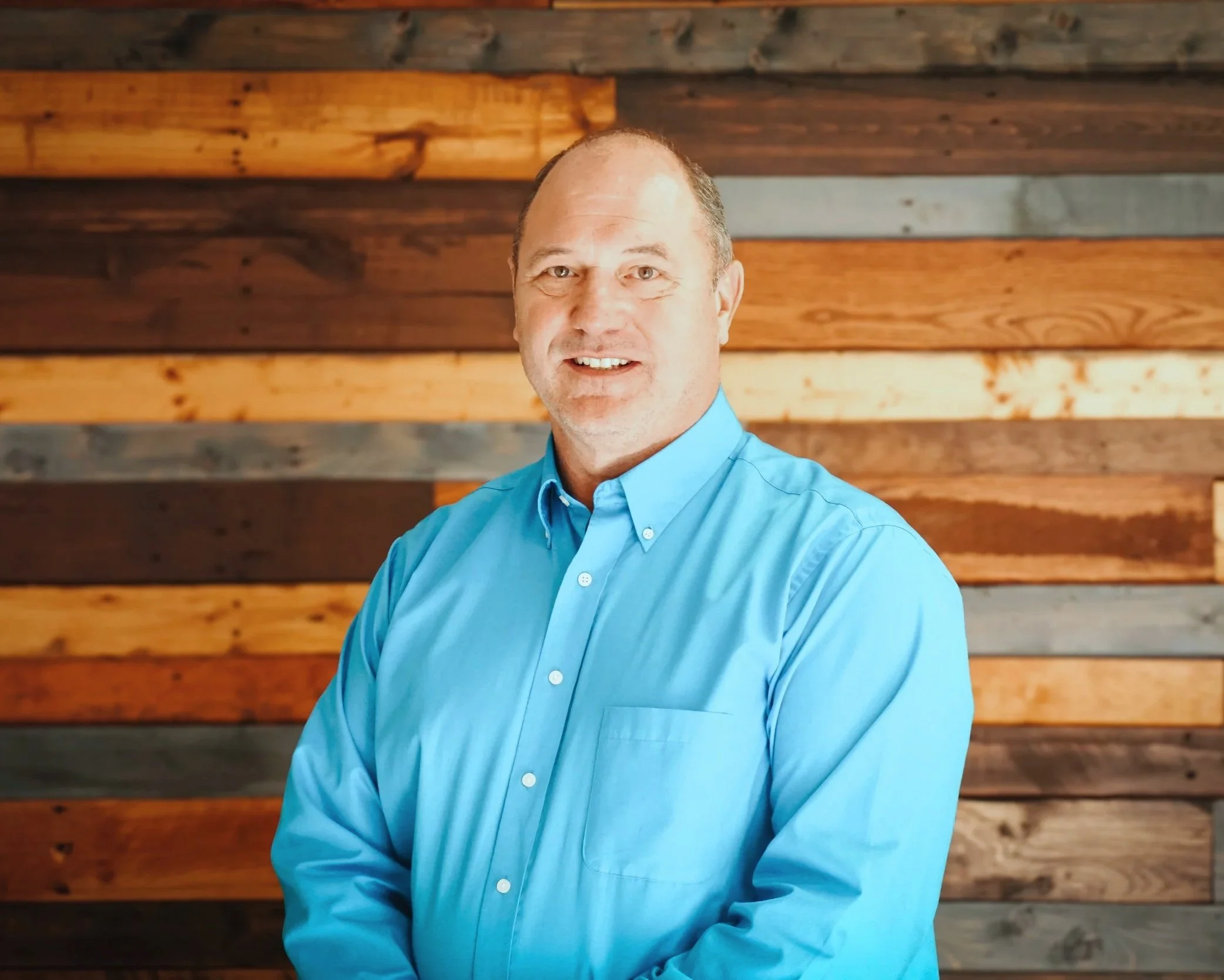 A man in a blue dress shirt standing in front of a wooden wall with various shades of brown and gray planks.