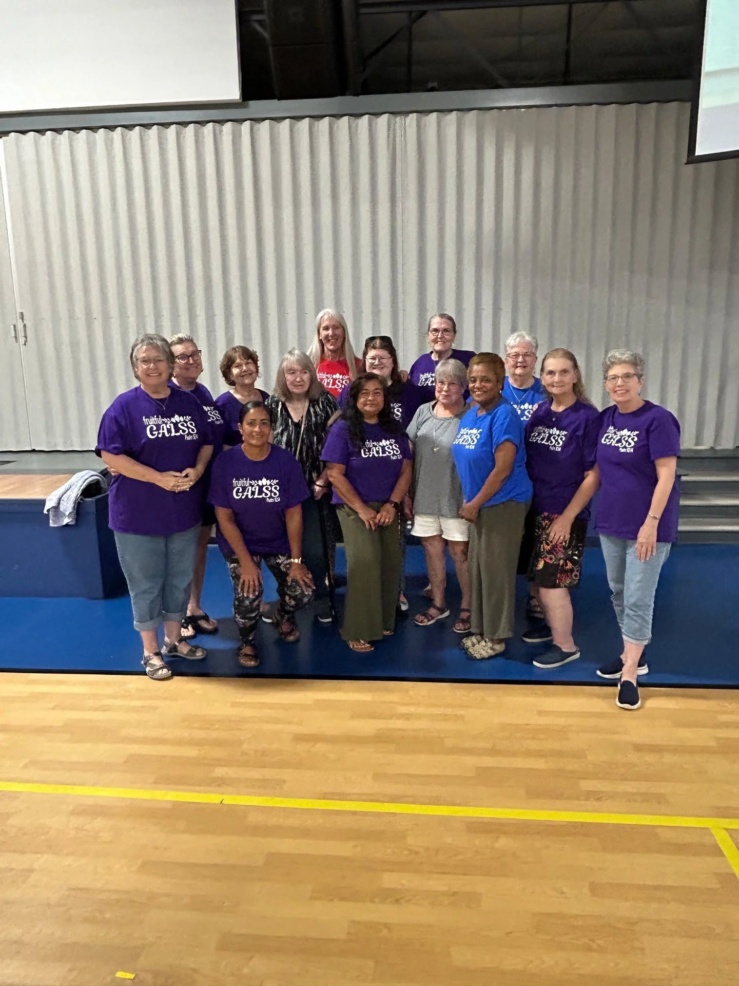 Group of fifteen women standing inside a gymnasium, some wearing purple shirts with white text, and some wearing other casual clothing, posing for a photo in front of a blue padded wall.
