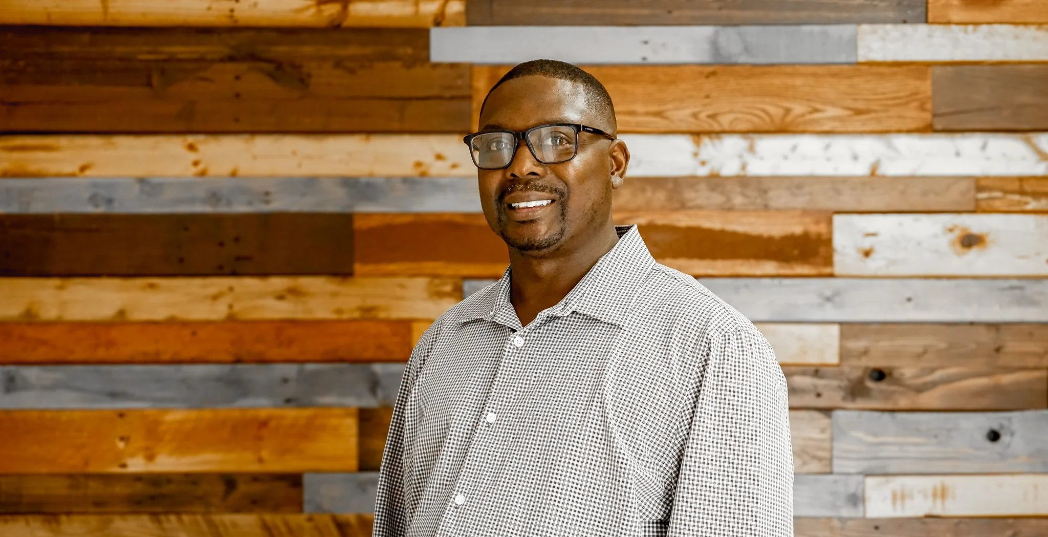 A man wearing glasses and a checked shirt standing in front of a wooden wall background.