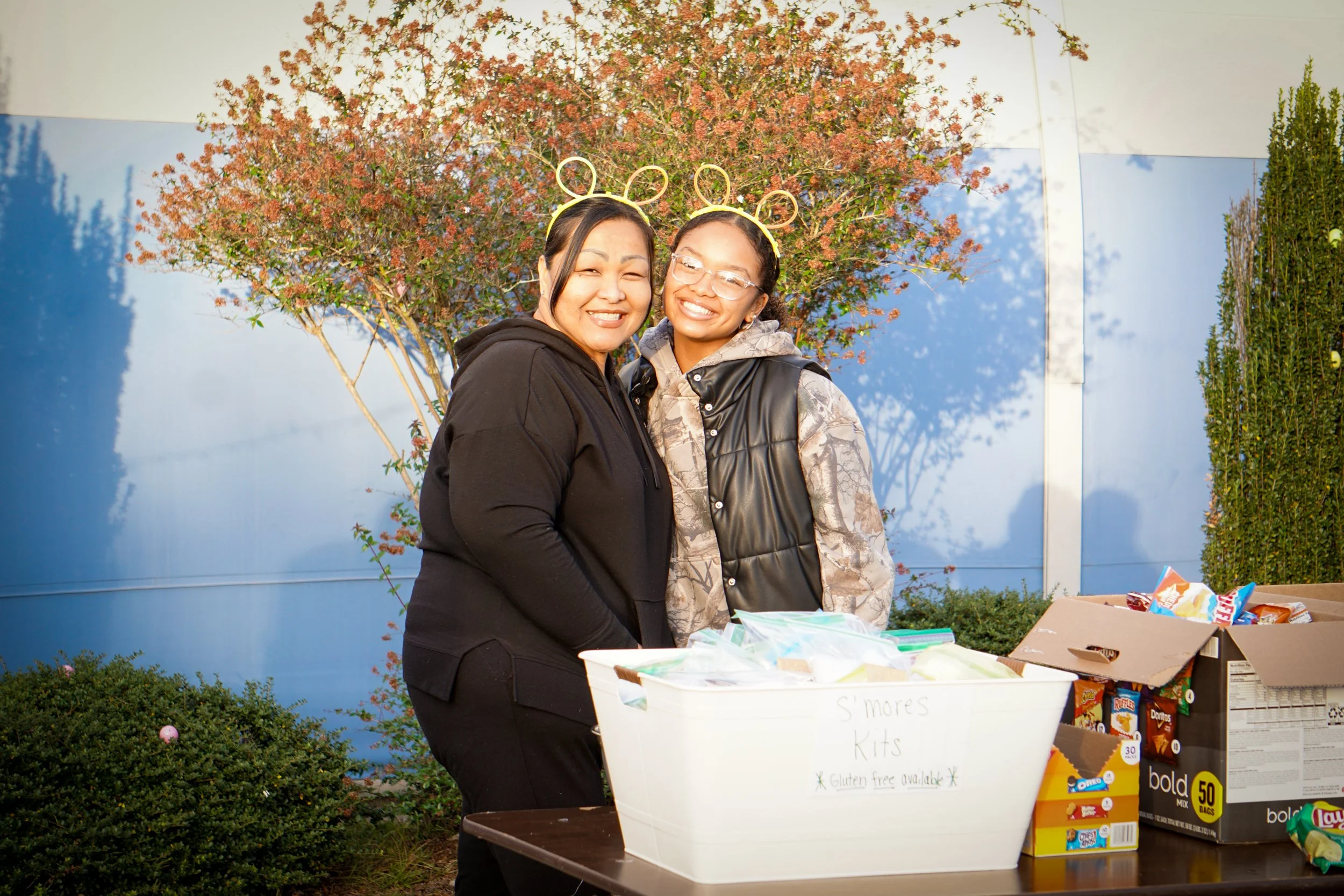 Two women wearing mouse ear headbands smiling at a fundraiser table with snacks, a blue wall and a flowering bush in the background.