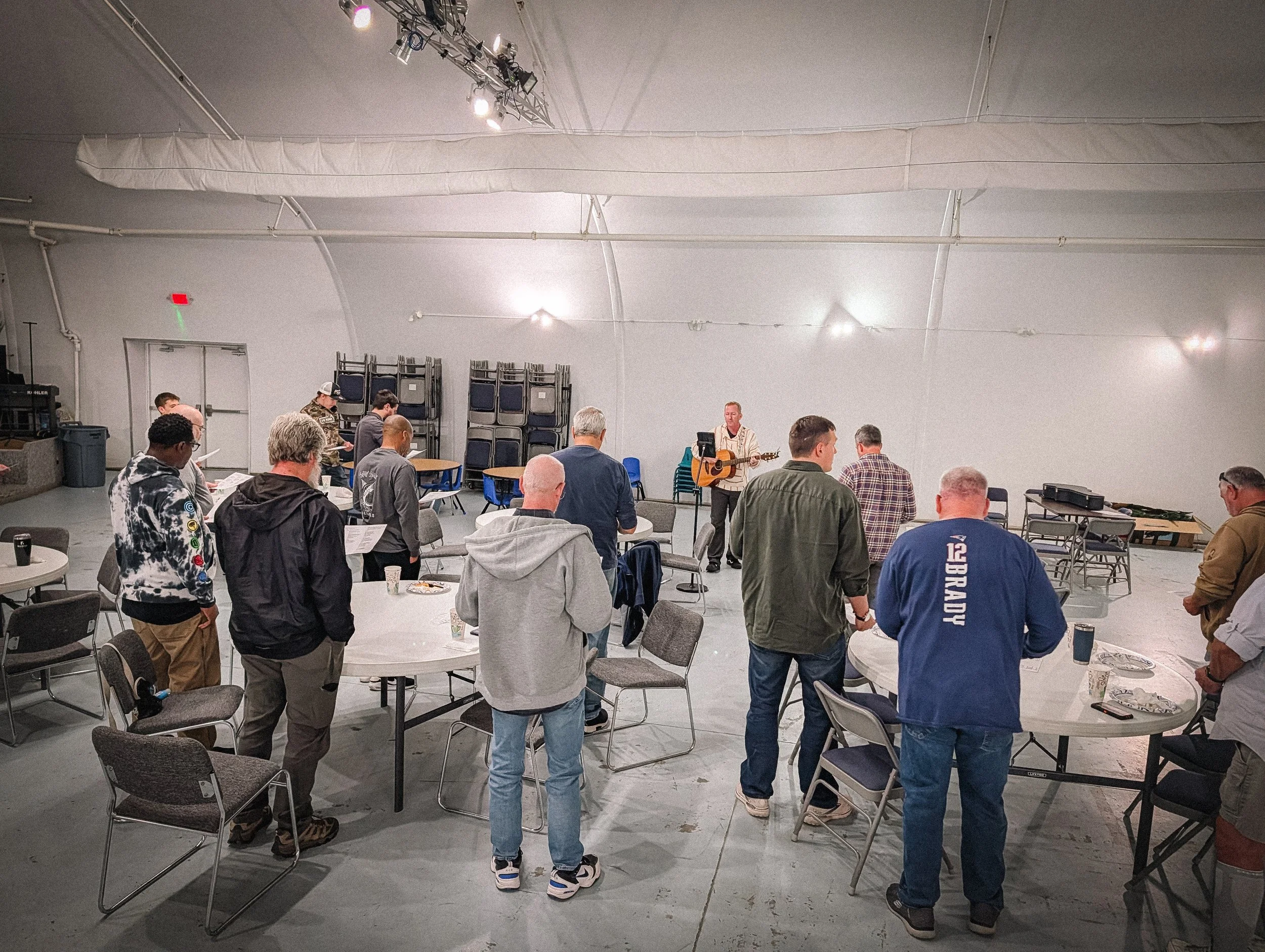 People standing and sitting at round tables in a large room, listening to a man playing guitar and singing on stage.