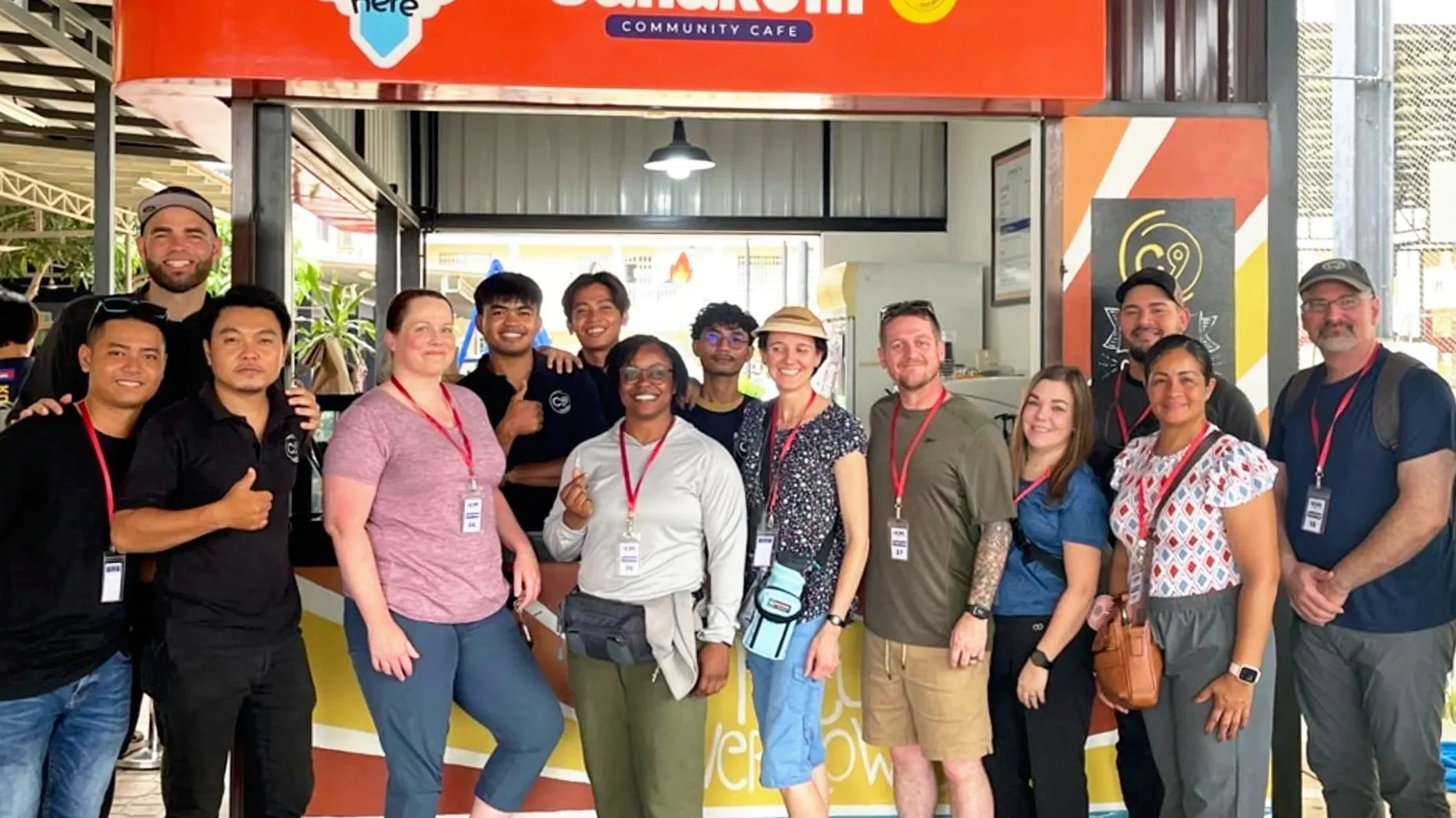 Group of fourteen diverse people smiling and posing for a photo at a community cafe, with some wearing badges and casual attire, inside a brightly lit area.