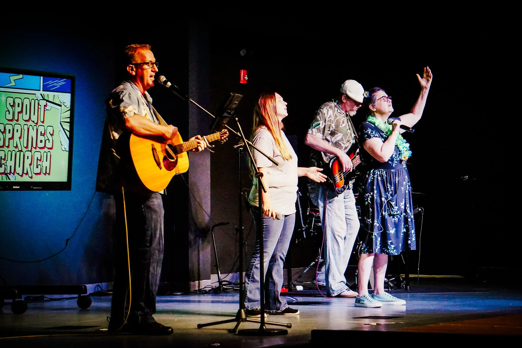 Group of five people performing on stage with musical instruments, including a man playing guitar, at Spout Springs Church.