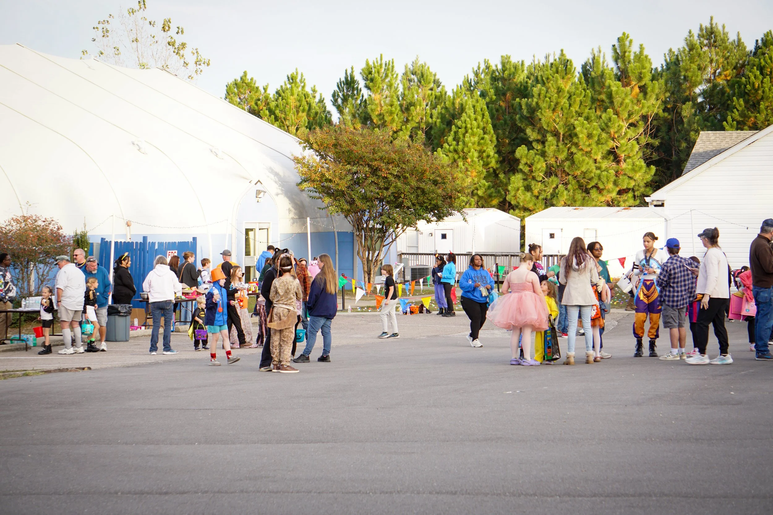 People gathered outdoors for a Halloween celebration, with some dressed in costumes like princesses and other characters. There are booths, decorations, and a string of colorful flags, with green trees and buildings in the background.