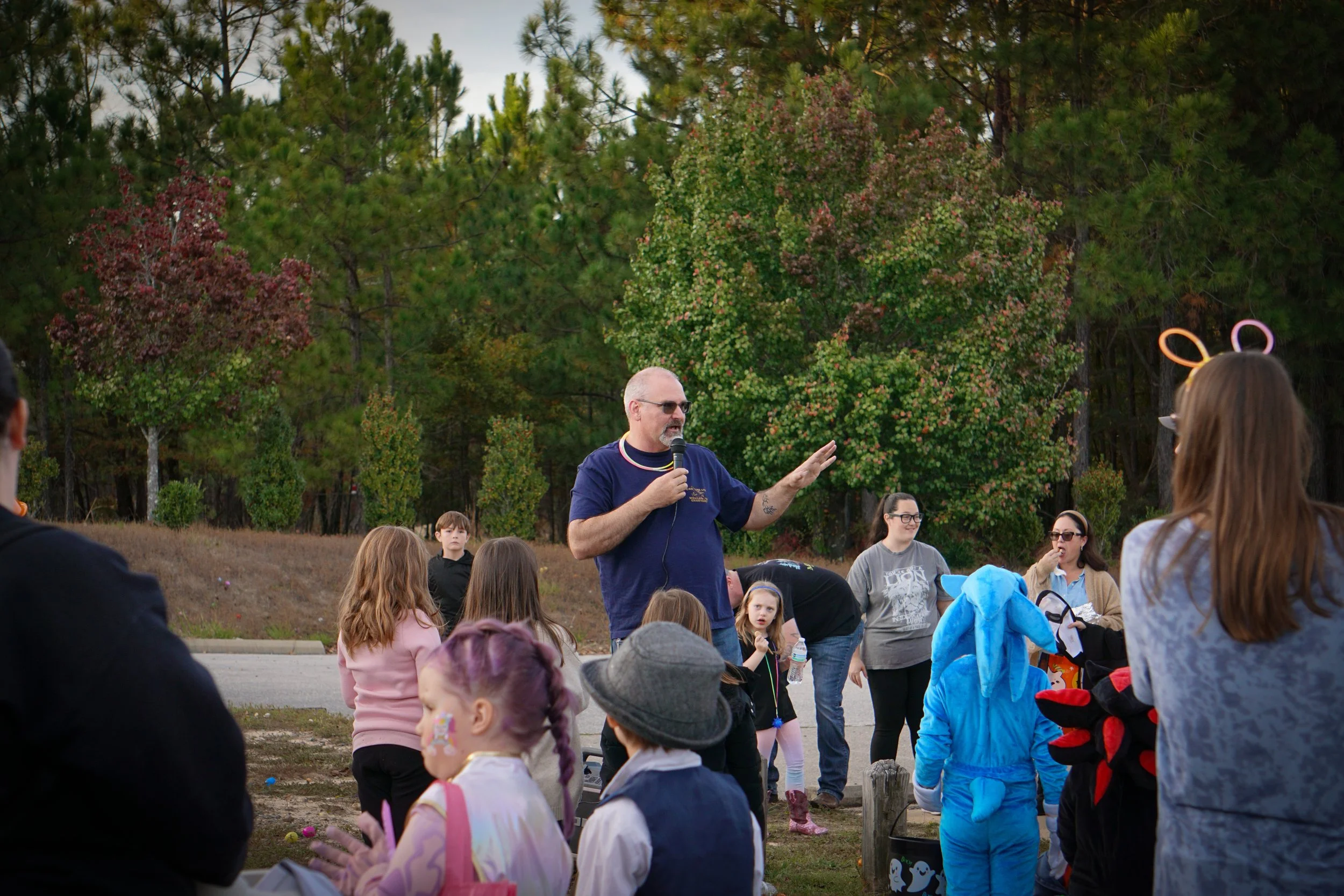 A man in sunglasses and a navy t-shirt addresses a crowd of children outdoors during daytime. The children are wearing costumes and jackets, with some facing the speaker and others looking around. Tall trees with green and reddish leaves are in the b