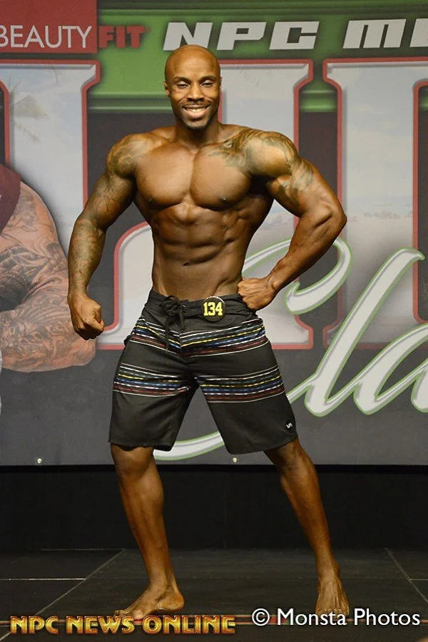 A muscular man is posing on stage at a bodybuilding competition, wearing black and striped shorts, with a number 134 on his shorts, smiling at the camera, with a colorful backdrop and text related to NPC fitness show.