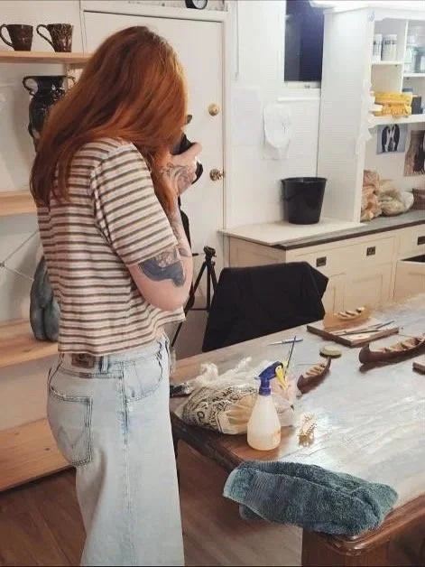 A woman with long red hair and tattoos, dressed in a striped shirt and light jeans, standing in a kitchen and looking at her phone.