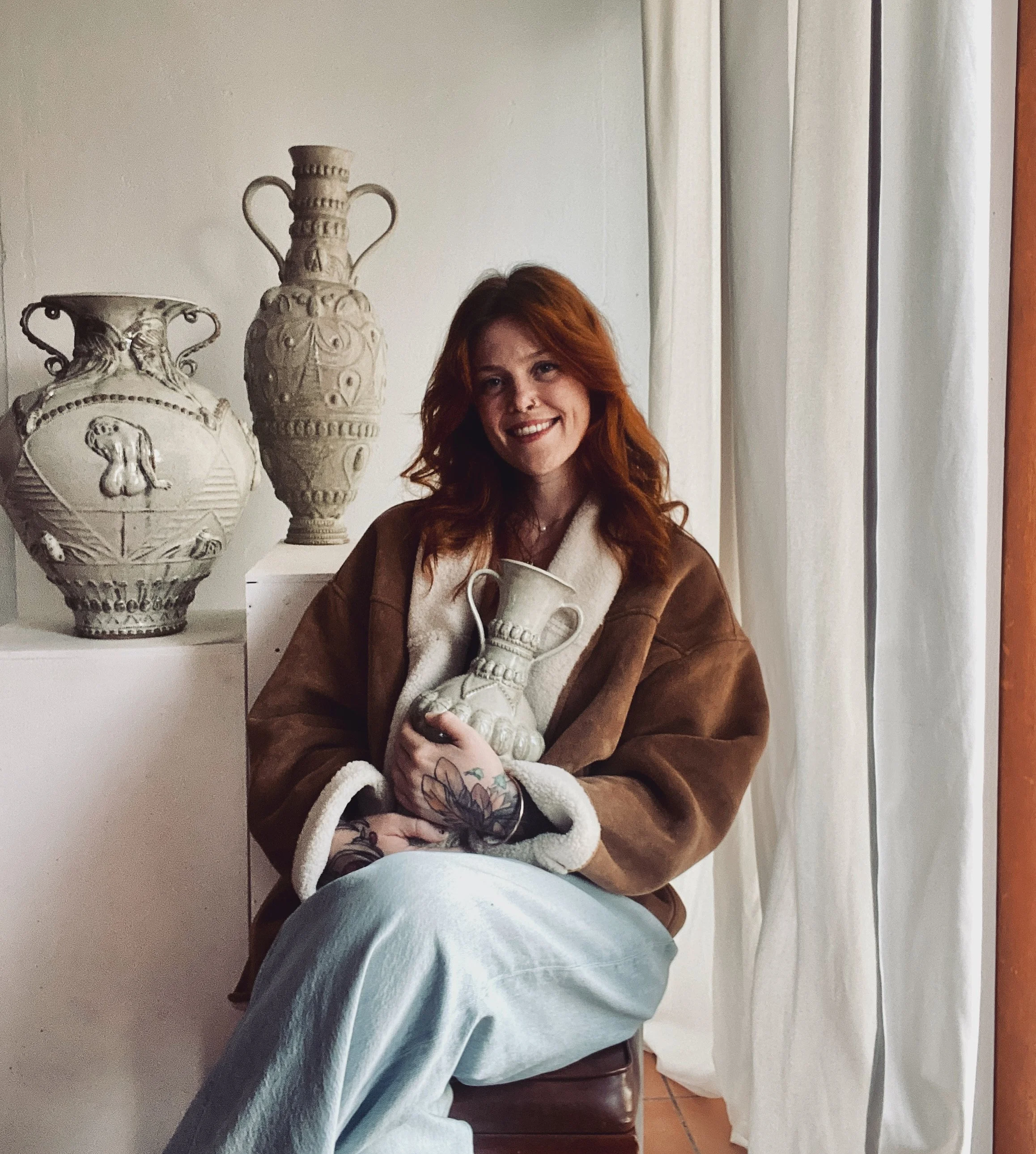 A woman with red hair and tattoos on her hand, sitting on a chair indoors, holding a ceramic vase, smiling at the camera. Behind her, ceramic vases are displayed on white pedestals, with cream-colored curtains beside her.