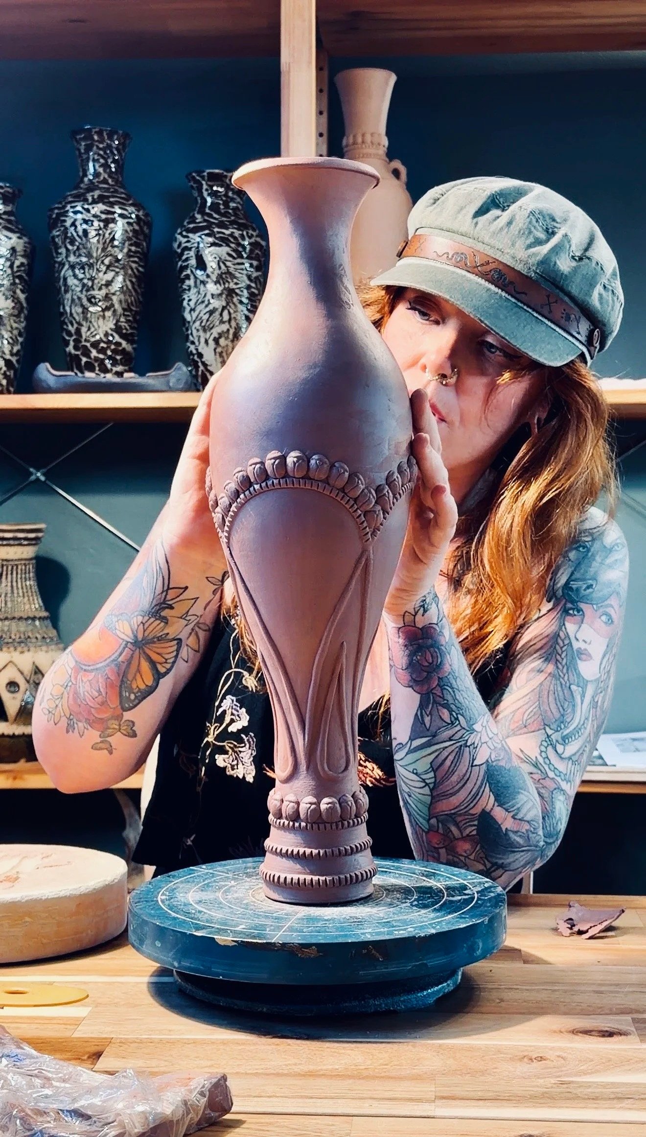 A woman with tattoos on her arms, wearing a cap, is carefully working on a tall, ornate clay vase in a pottery studio.