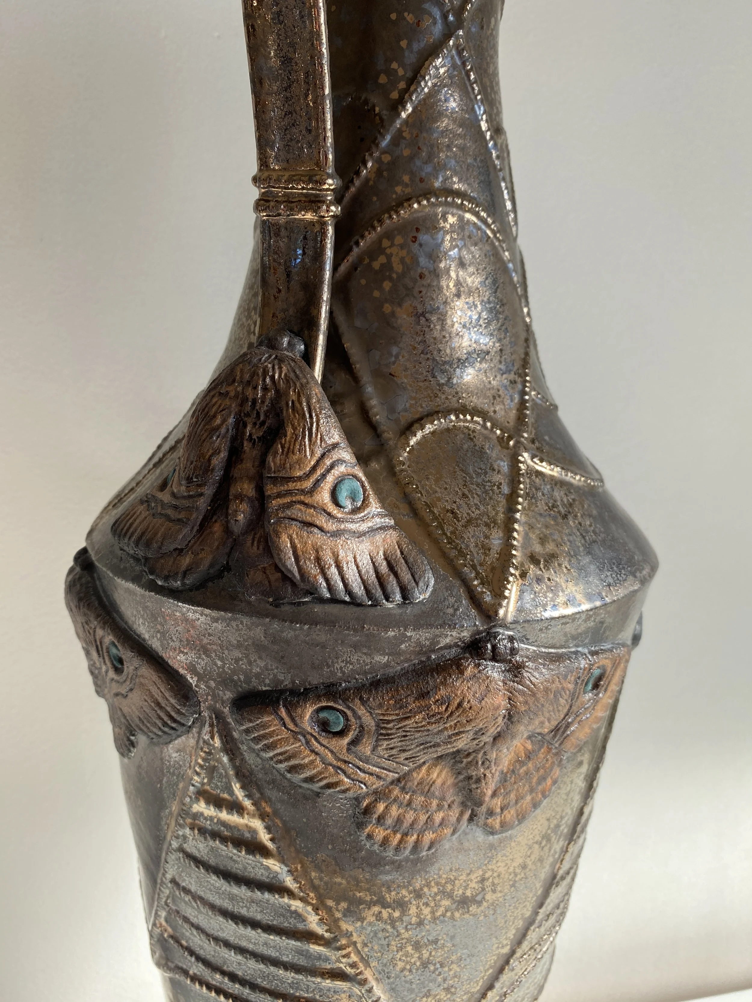 Close-up of a metal decorative vase with intricate fish and leaf motifs, including textured fish scales and eye details, in a bronze finish.