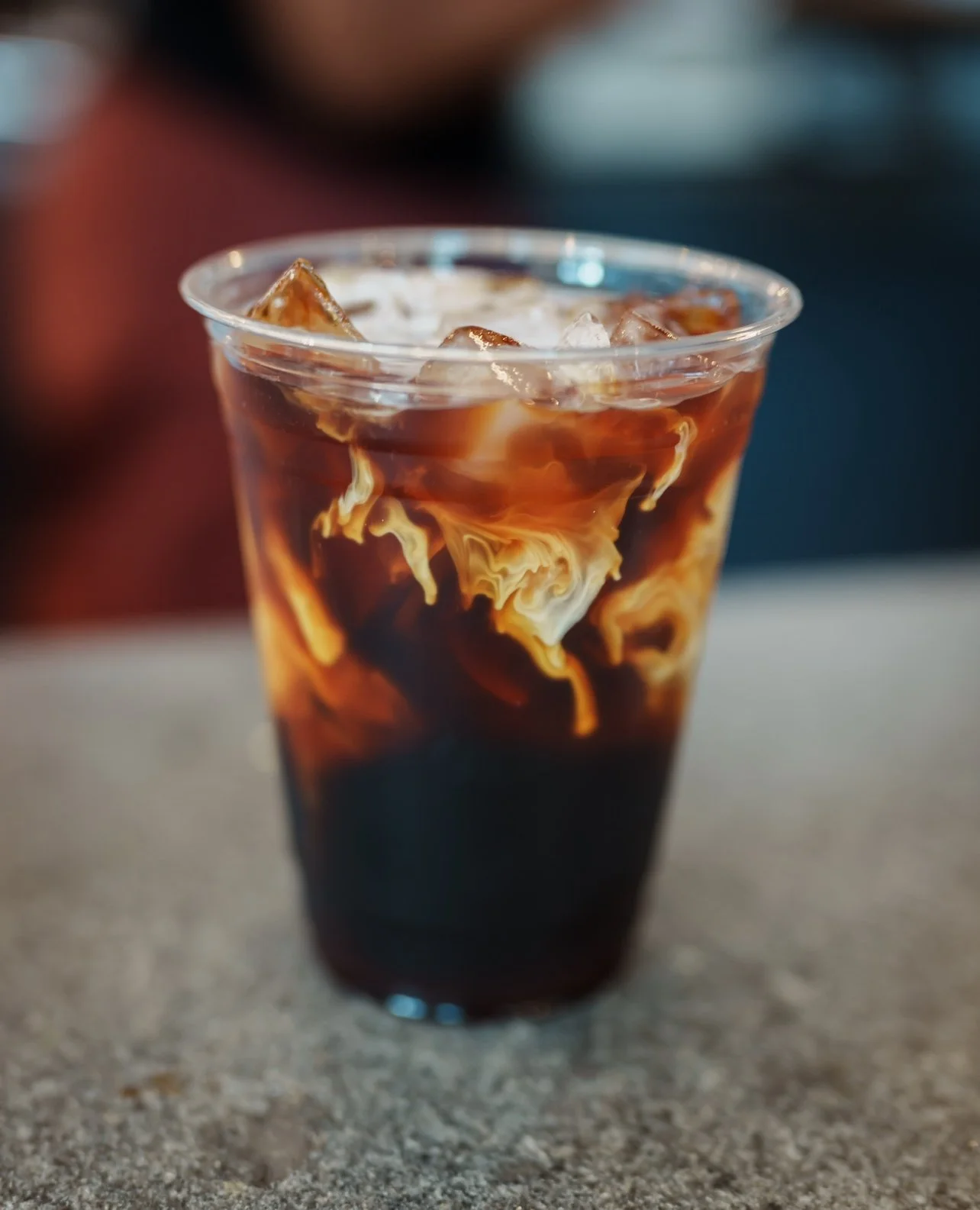 A clear plastic cup filled with iced coffee, with cream swirling into the dark coffee creating a marbled effect.