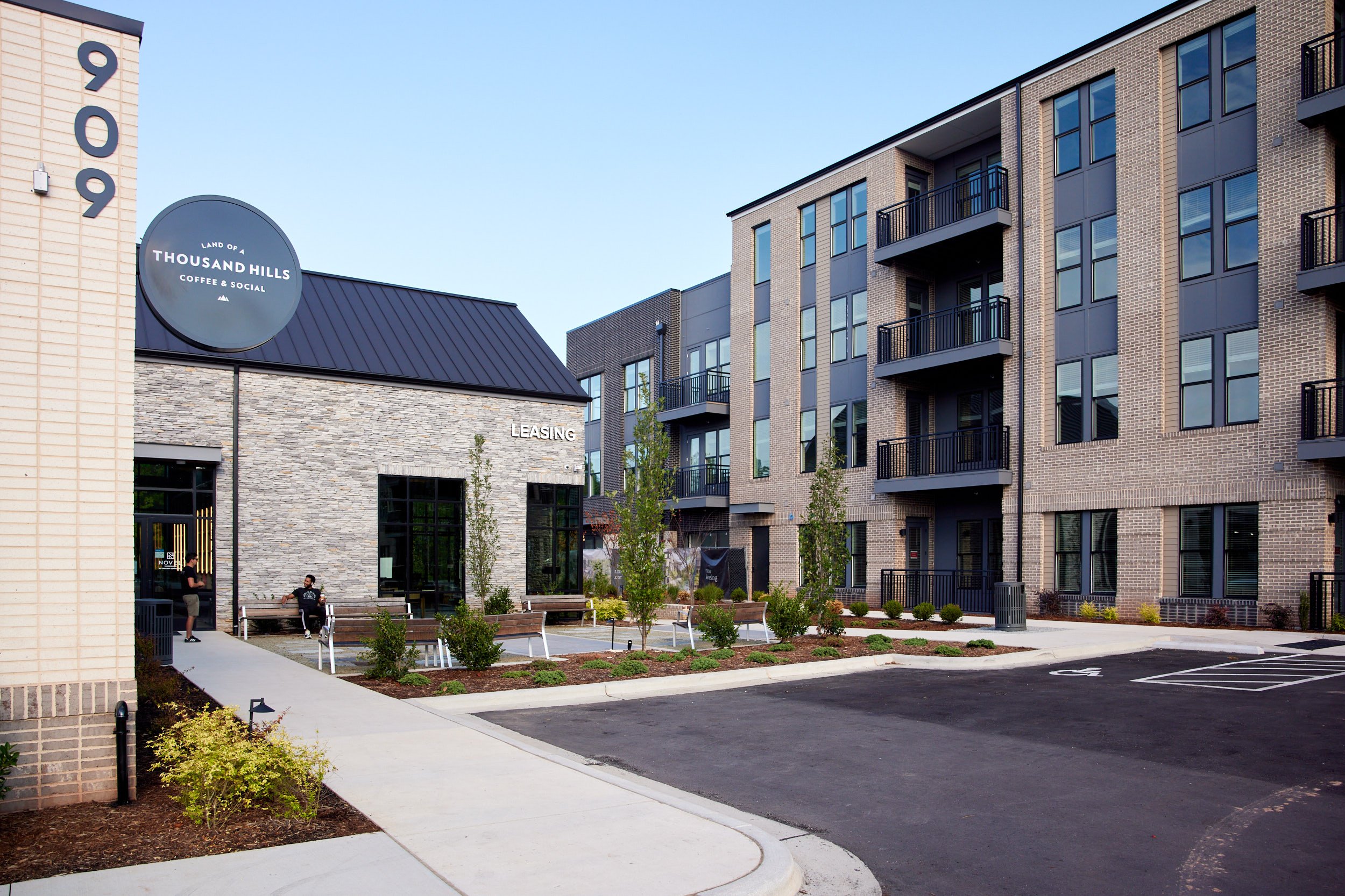 Modern apartment complex with a landscaped courtyard, parking lot, and a coffee shop called 'Land of a Thousand Hills Coffee & Social' in the foreground.