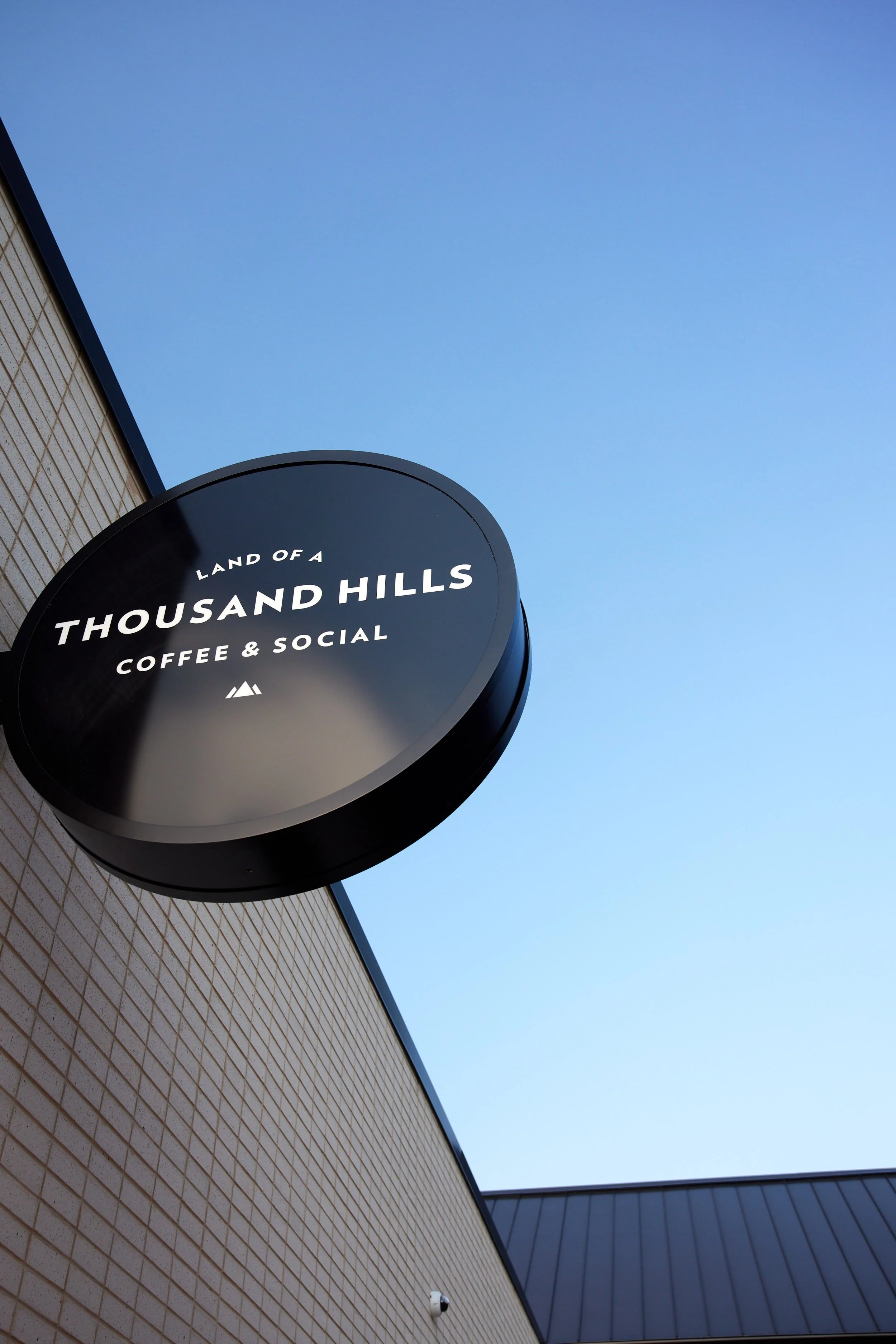 Black circular sign on beige brick wall with white text, reads "Land of a Thousand Hills Coffee & Social" against a clear blue sky.