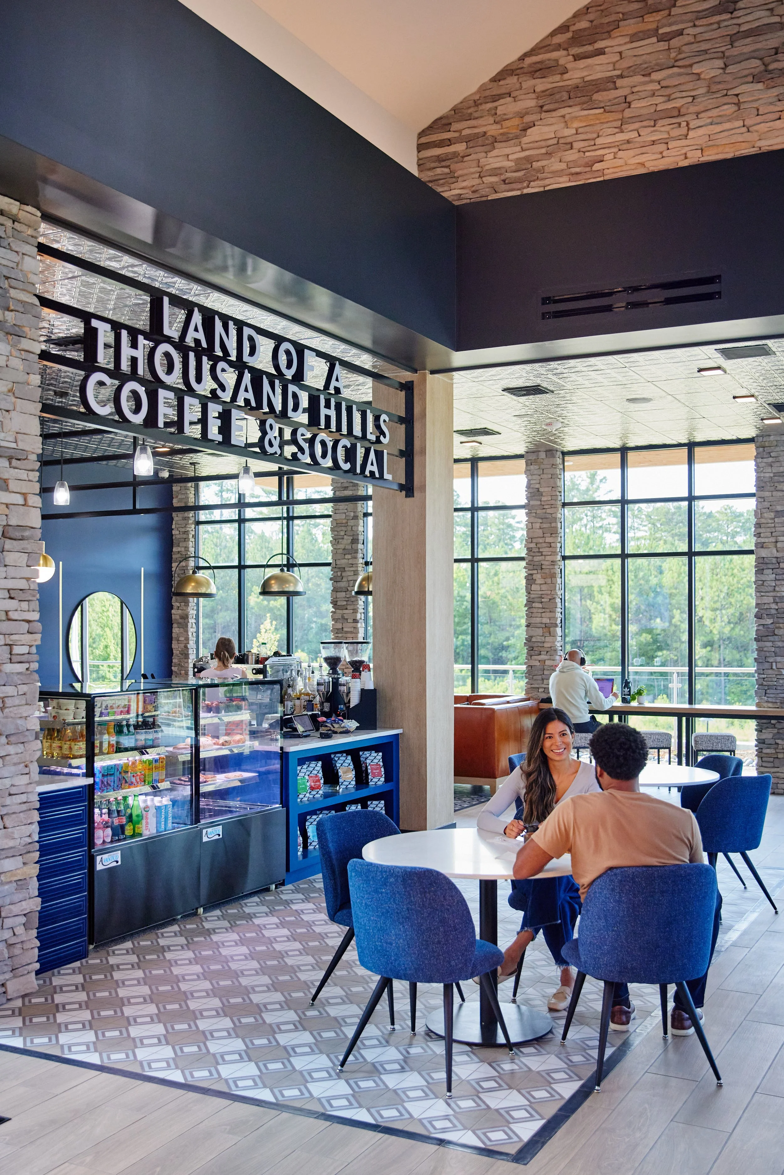 Modern coffee shop with large windows, stone wall accents, blue chairs, and a sign that reads "Land of a Thousand Hills Coffee & Social." Two people are seated at a white table, smiling and conversing, while a barista prepares drinks behind the count